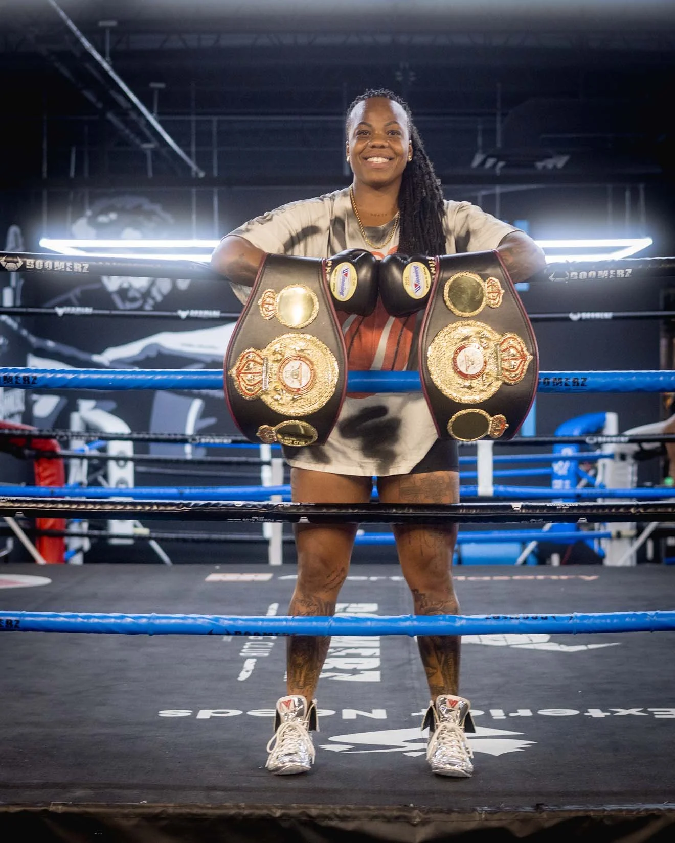 A female boxer standing inside a boxing ring, smiling with boxing belts draped over the top rope in front of her, in a gym with boxing-themed decor.
