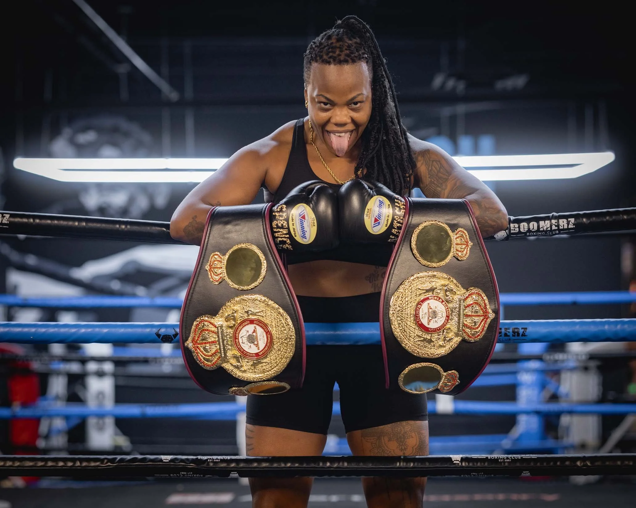 A female boxer in black athletic wear sticking out her tongue, resting her arms on boxing championship belts in a boxing ring.