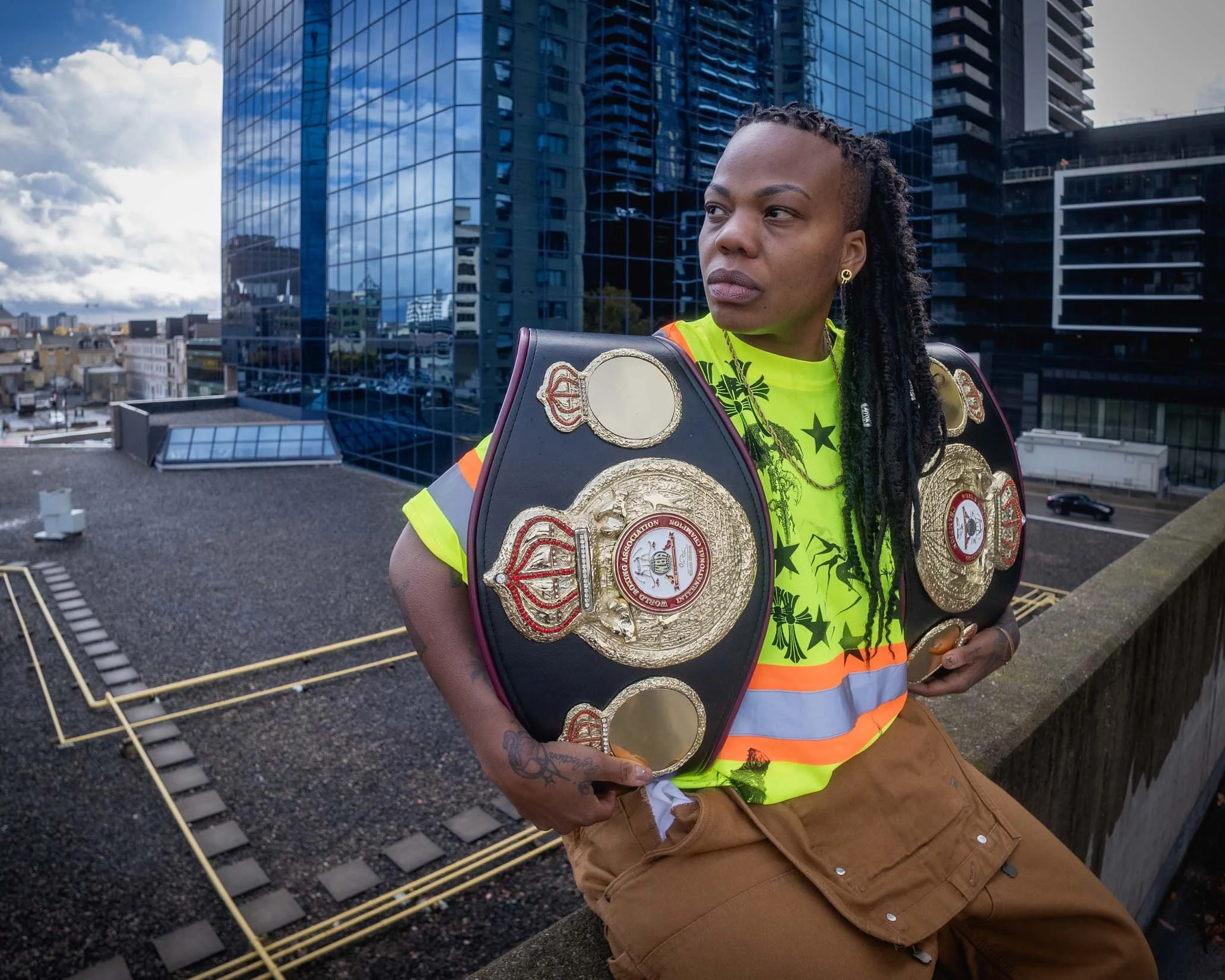 A woman with braided hair, wearing a neon yellow safety vest with orange reflective stripes, stands on a rooftop overlooking a cityscape. She has multiple championship belts draped over her shoulder and arm, and is dressed in tan pants.