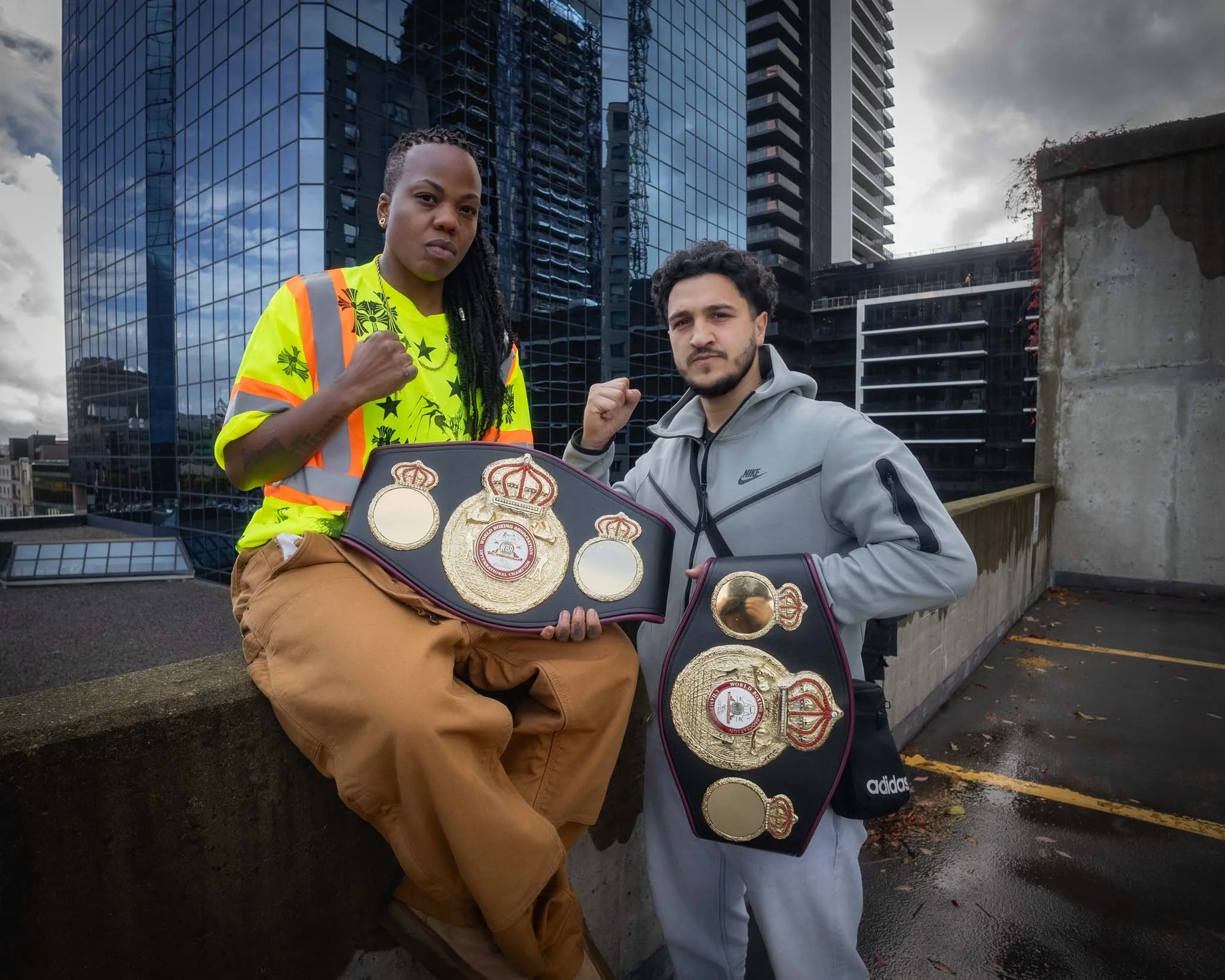 Two individuals standing outdoors in an urban area with modern glass buildings behind them. They are posing with fists raised and wearing boxing championship belts. One individual is wearing a yellow high-visibility safety vest and brown pants, while