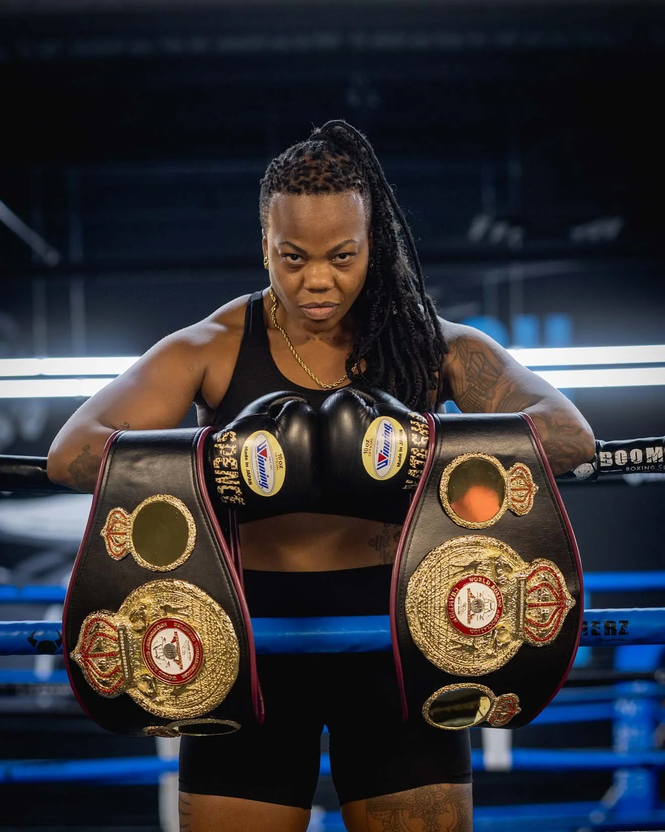 A female boxer with braided hair and tattoos, wearing a black sports bra and shorts, standing inside a boxing ring with championship belts draped over the ropes in front of her, looking determined.