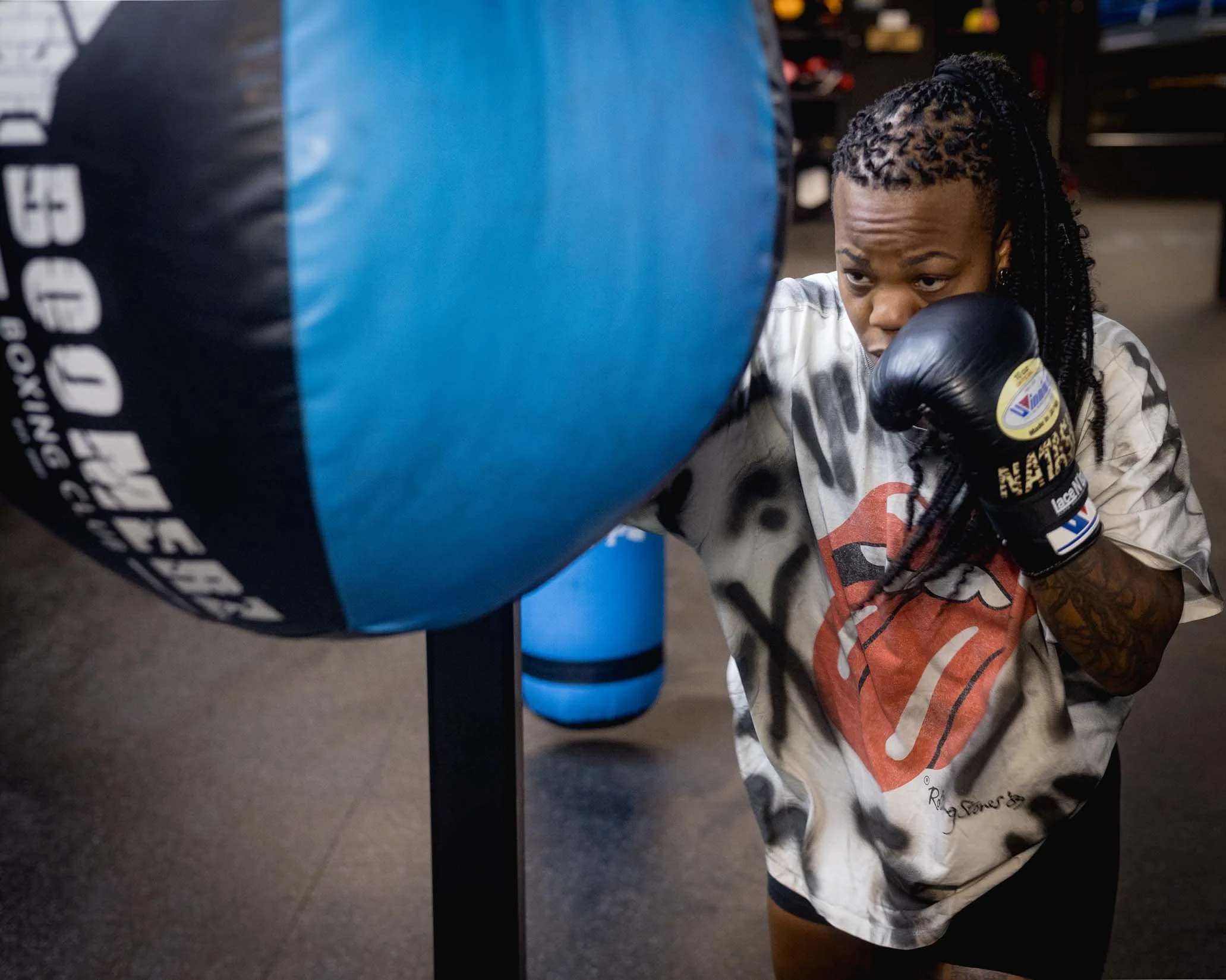 A young girl with braided hair practicing boxing with a black punching bag in a gym.