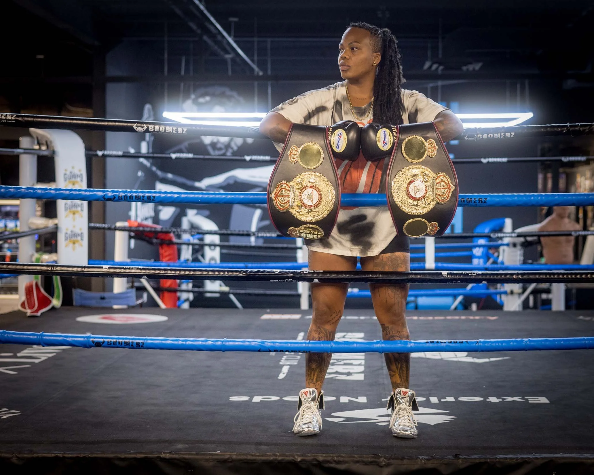 A female boxer stands inside a boxing ring with championship belts hanging from her neck, her arms resting on the ropes, looking to the side, in a modern gym with dark lighting and neon lights.
