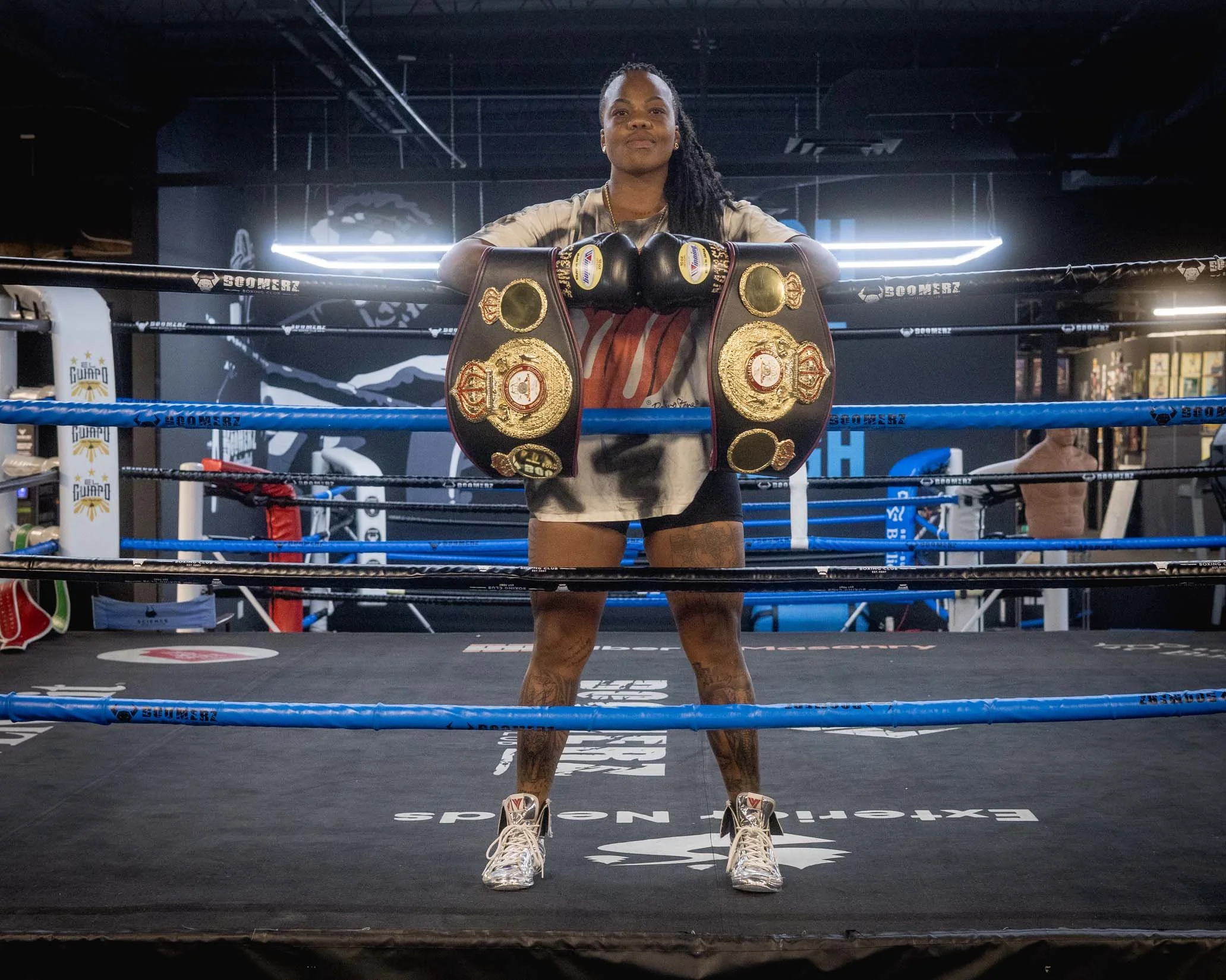 A female boxer standing in a boxing ring with her arms resting on the top rope, wearing boxing gloves and championship belts around her neck and waist, inside a gym with dark lighting and boxing equipment.