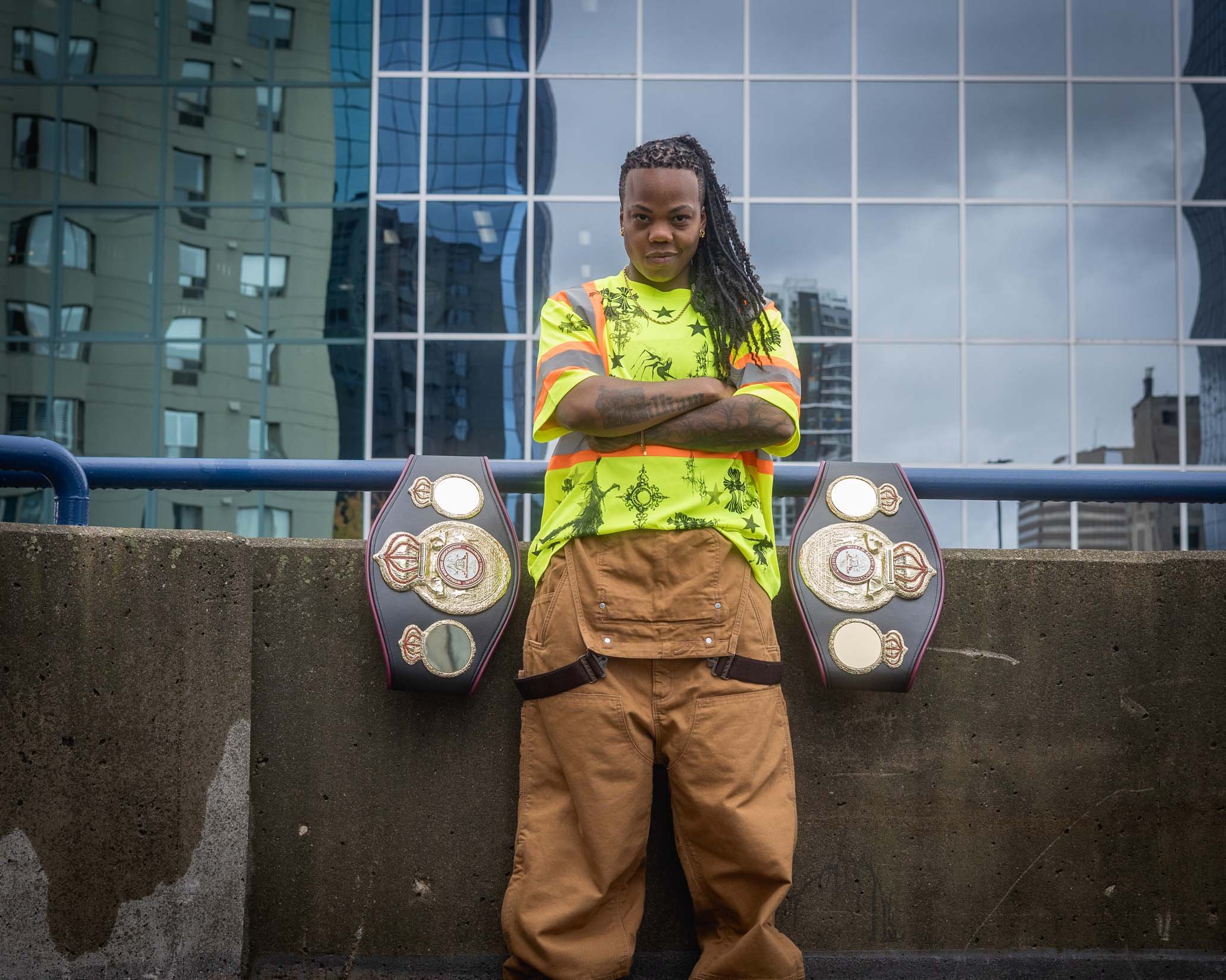 A person with dark skin and dreadlocked hair standing with arms crossed in front of a modern glass building, wearing a bright yellow-green graphic t-shirt and khaki cargo pants, flanked by championship belts on a ledge.