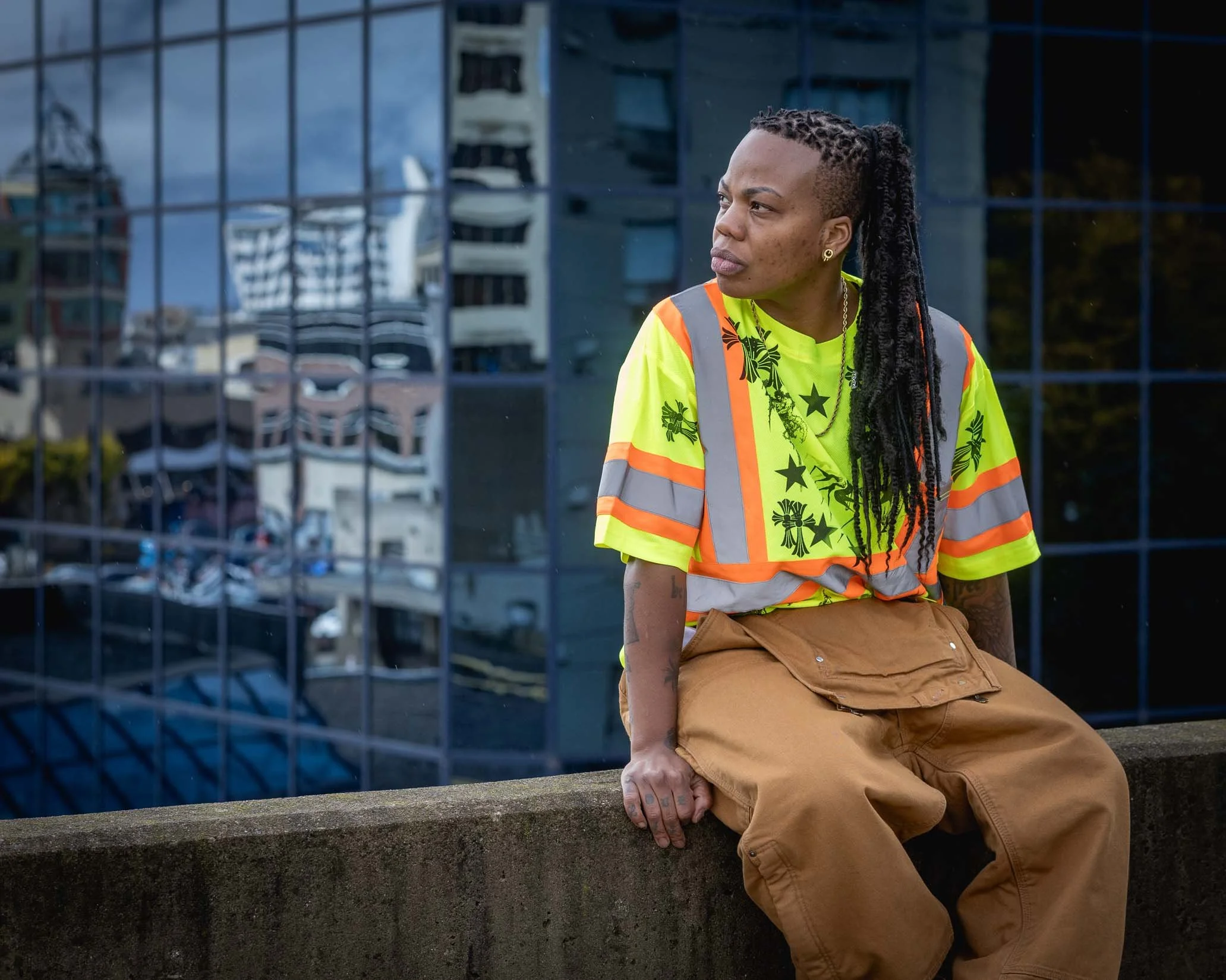 A person with long dreadlocks, wearing earrings, a bright yellow shirt with black stars and green designs, a safety vest, and tan pants, sitting on a concrete ledge in an urban setting with glass buildings reflected in the background.