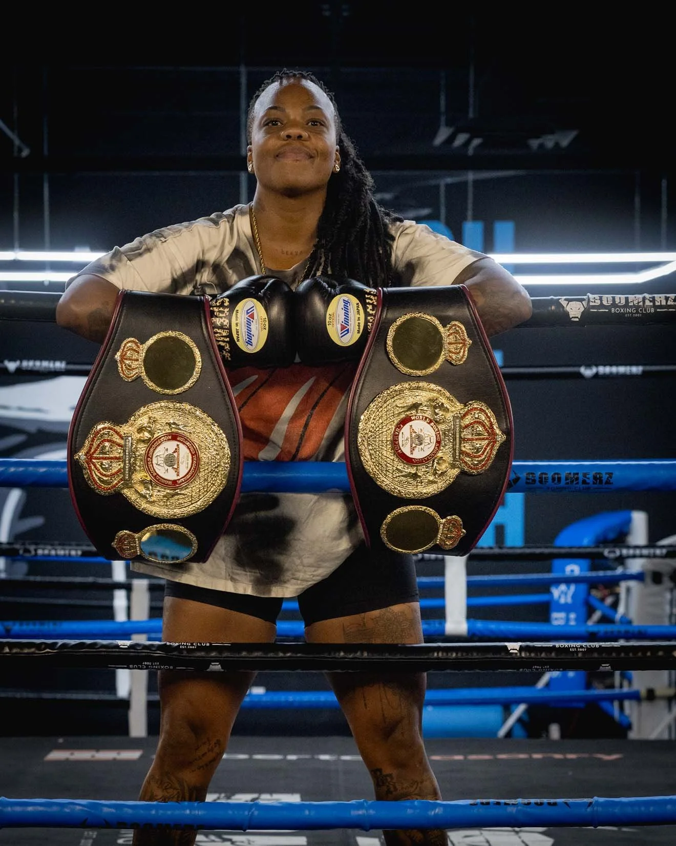 Smiling female boxer with long braids in a boxing ring, resting her arms on the ropes, with two championship belts draped over her forearms.