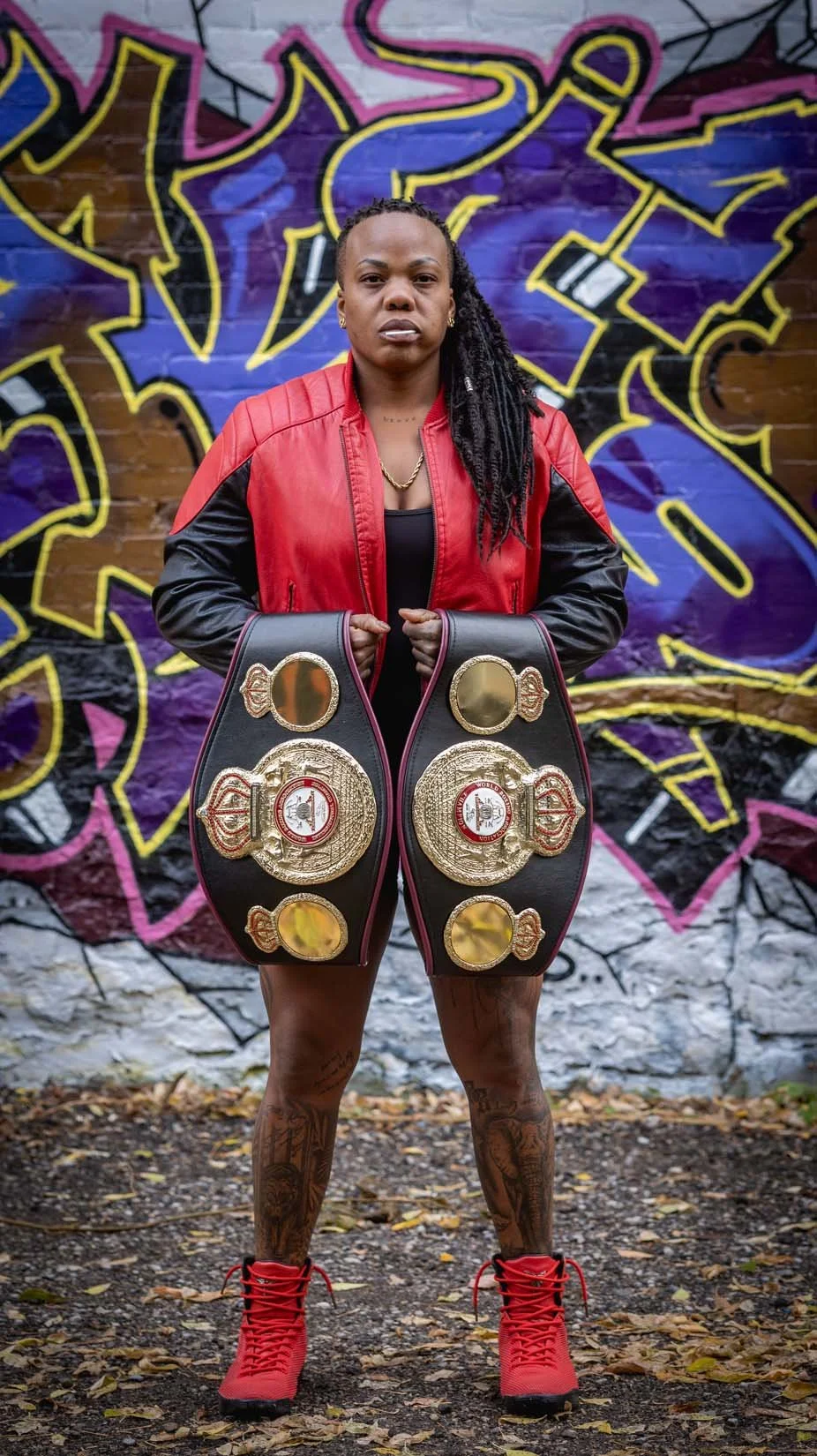 A woman with braids wearing a red and black leather jacket holds two championships belts in front of a colorful graffiti wall.