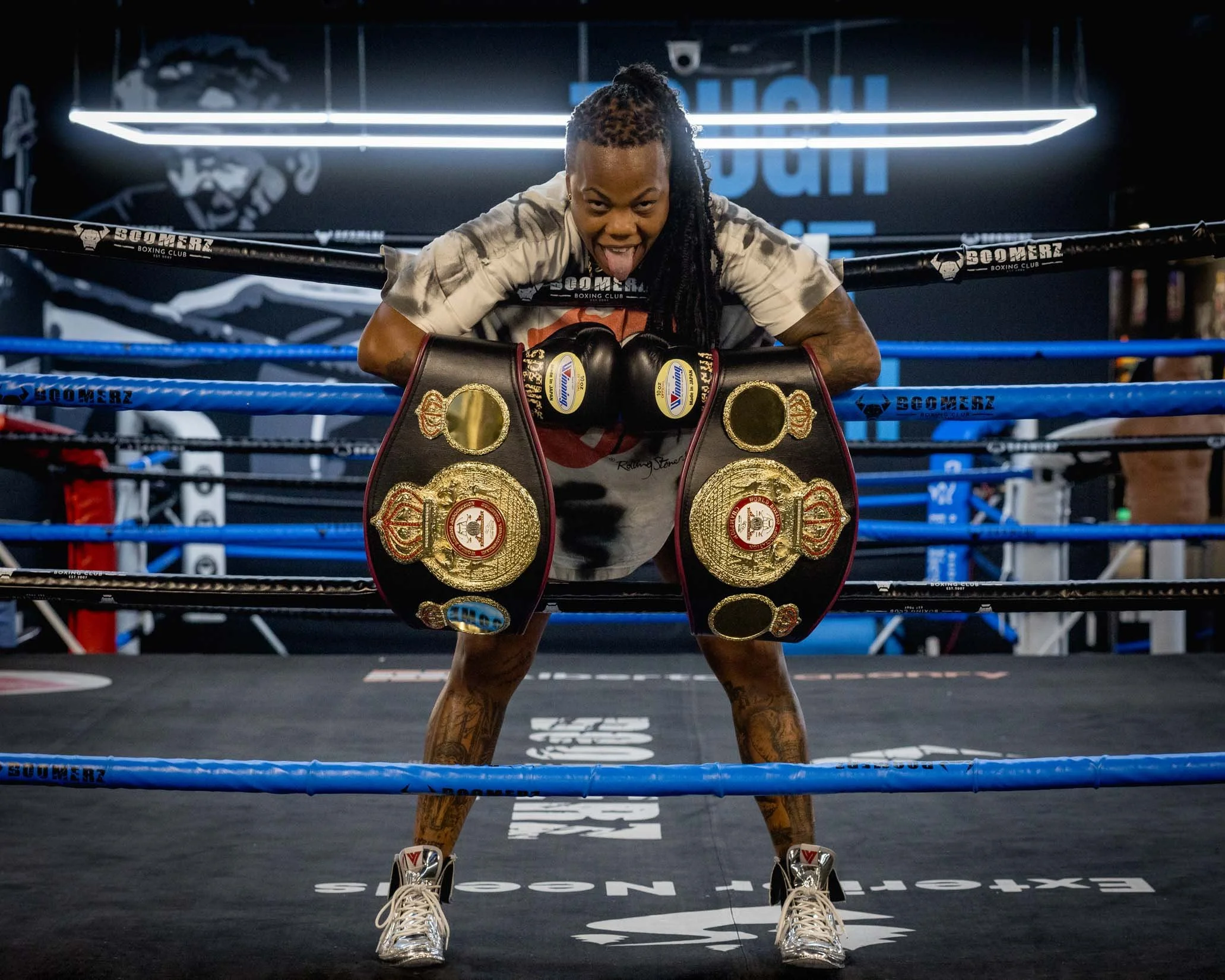 A female boxer in boxing gloves and championship belts leaning on the ropes of a boxing ring, tongue out, in a gym with dark background and bright lighting.