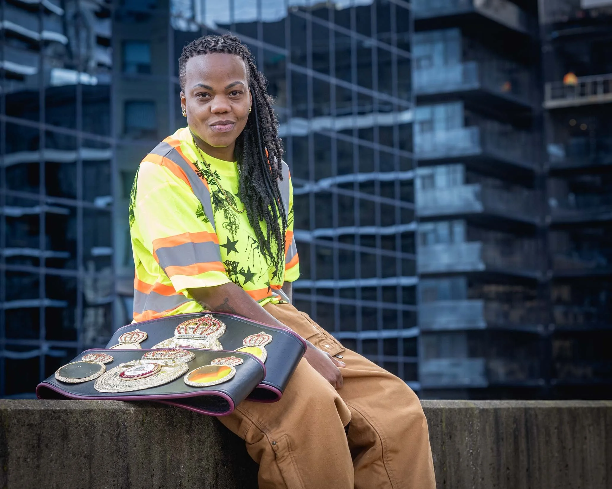 A woman with dreadlocks wearing a yellow safety vest and earrings sitting outdoors, holding a tray of embroidered patches with various designs, in front of a modern glass building.
