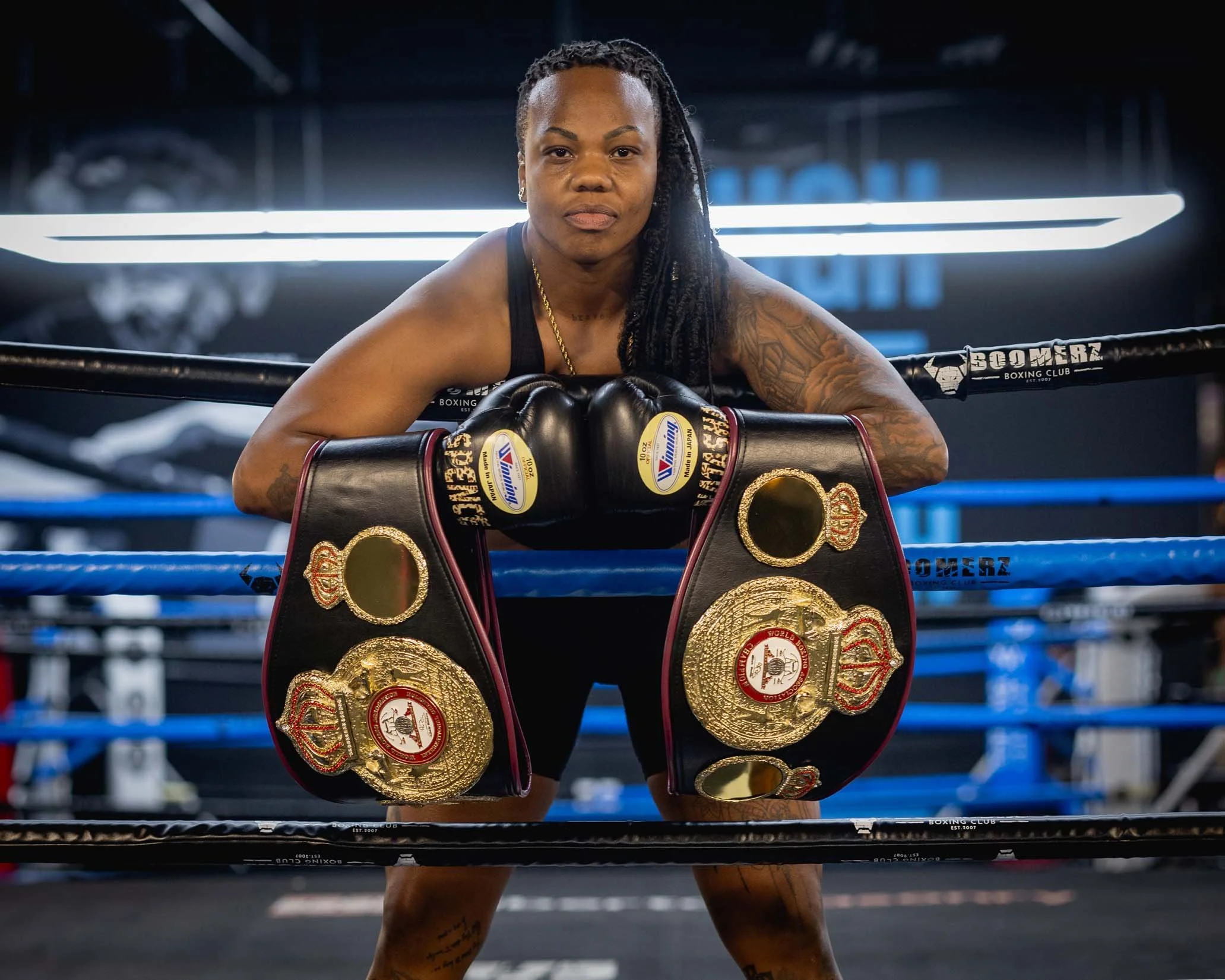 A female boxer with long, braided hair and tattoos, leaning on boxing gloves in a boxing ring with championship belts around her shoulders, inside a gym.