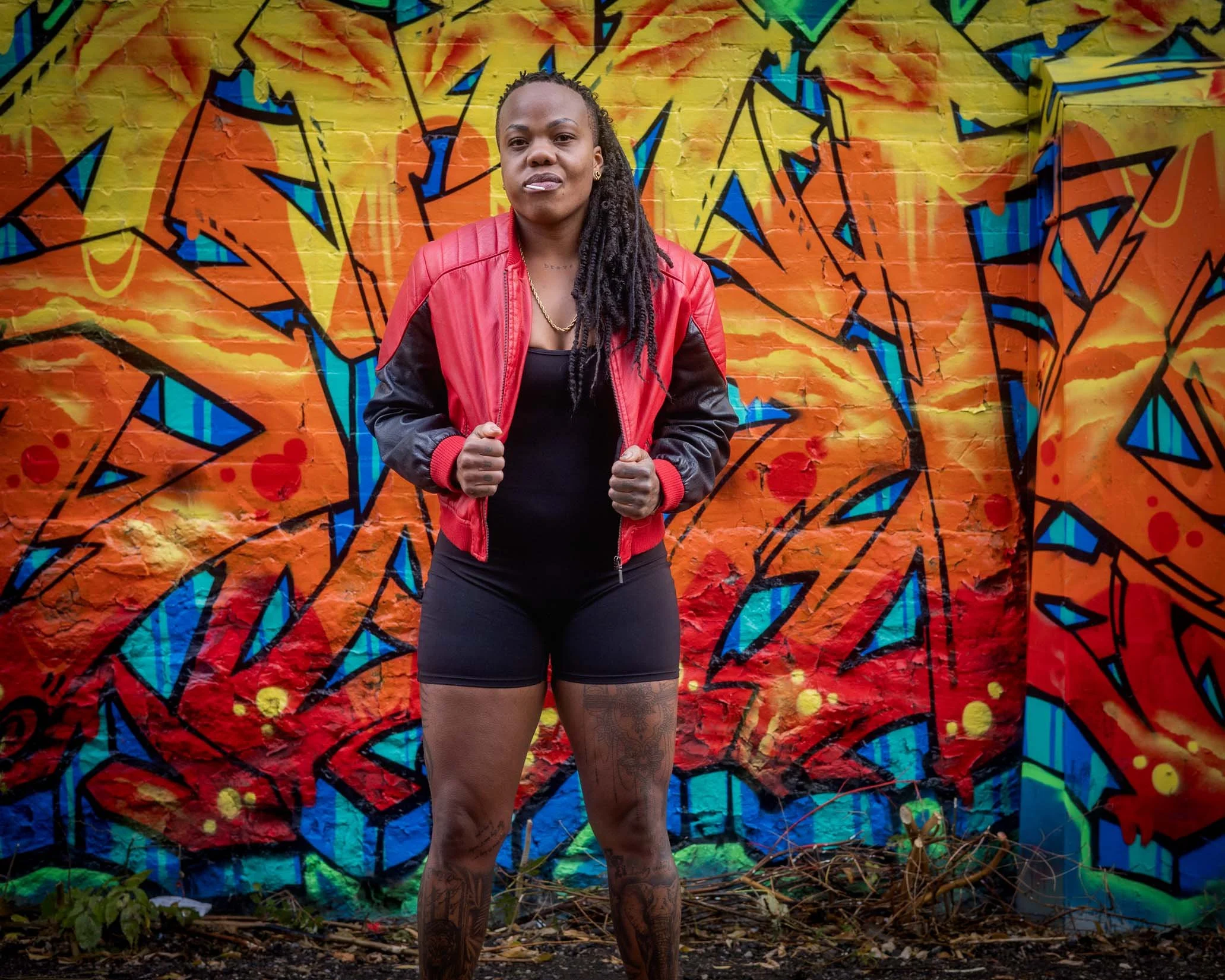 A woman with long dreadlocks stands confidently in front of a vivid, colorful graffiti mural on a brick wall. She is wearing a red and black jacket, a black top, and has tattoos on her legs and arms.