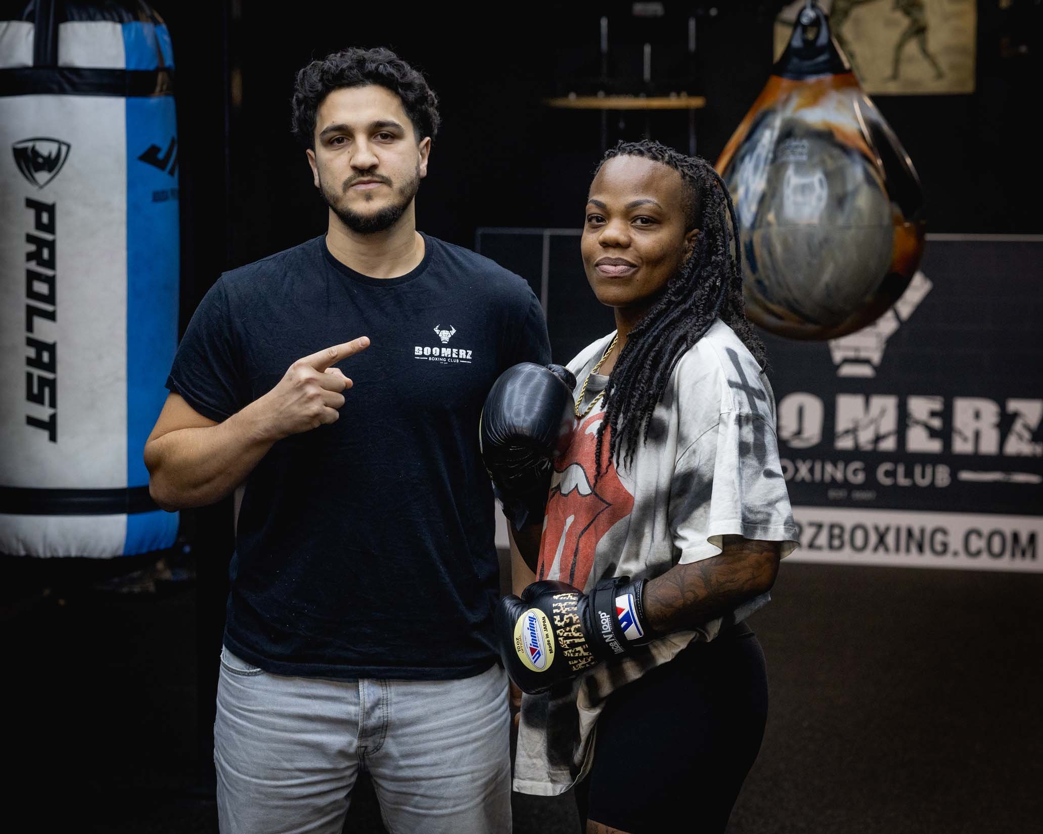 A man and woman in boxing gear standing together in a gym. The man has dark, curly hair and facial hair, and is wearing a black t-shirt with a logo that says 'BOOMERZ BOXING CLUB.' The woman has long dreadlocks, a confident expression, and is dressed