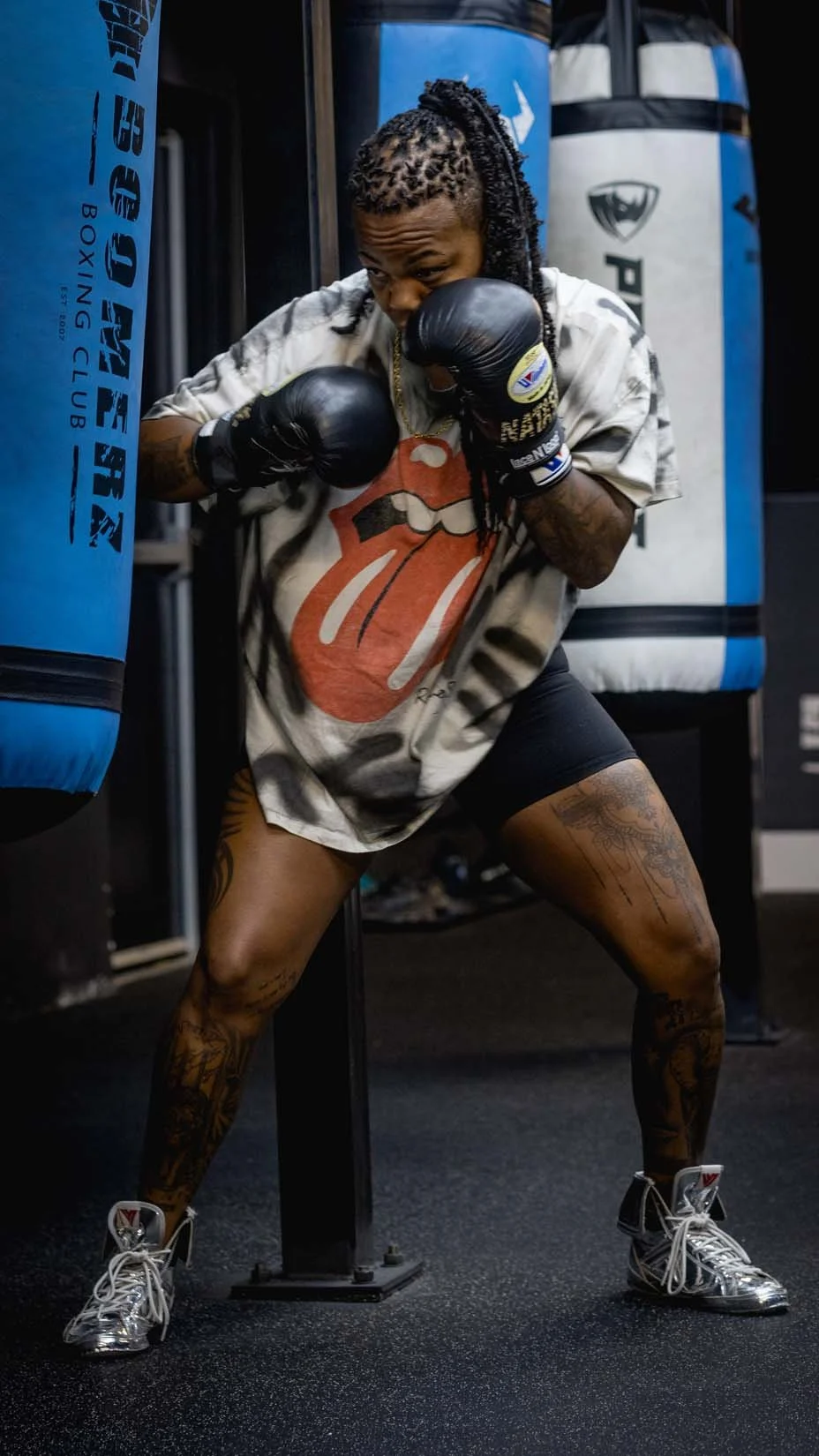 A woman wearing boxing gloves and a Rolling Stones t-shirt practices boxing on a punching bag at a gym.