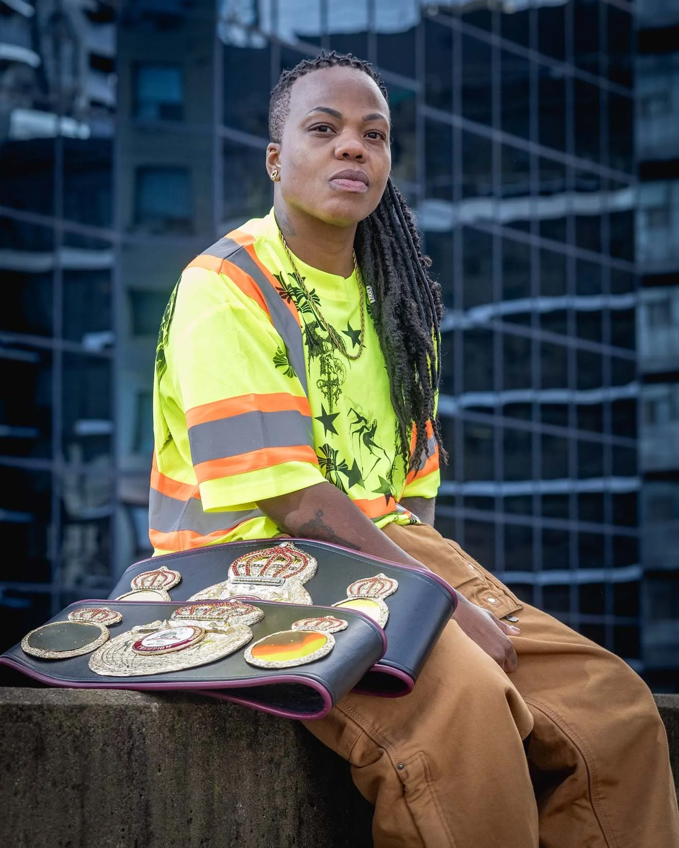 A person with long dreadlocks wearing a bright yellow safety vest with orange and gray stripes, sitting outdoors in front of a modern glass building, with boxing championship belts placed on a suitcase or bag in front.