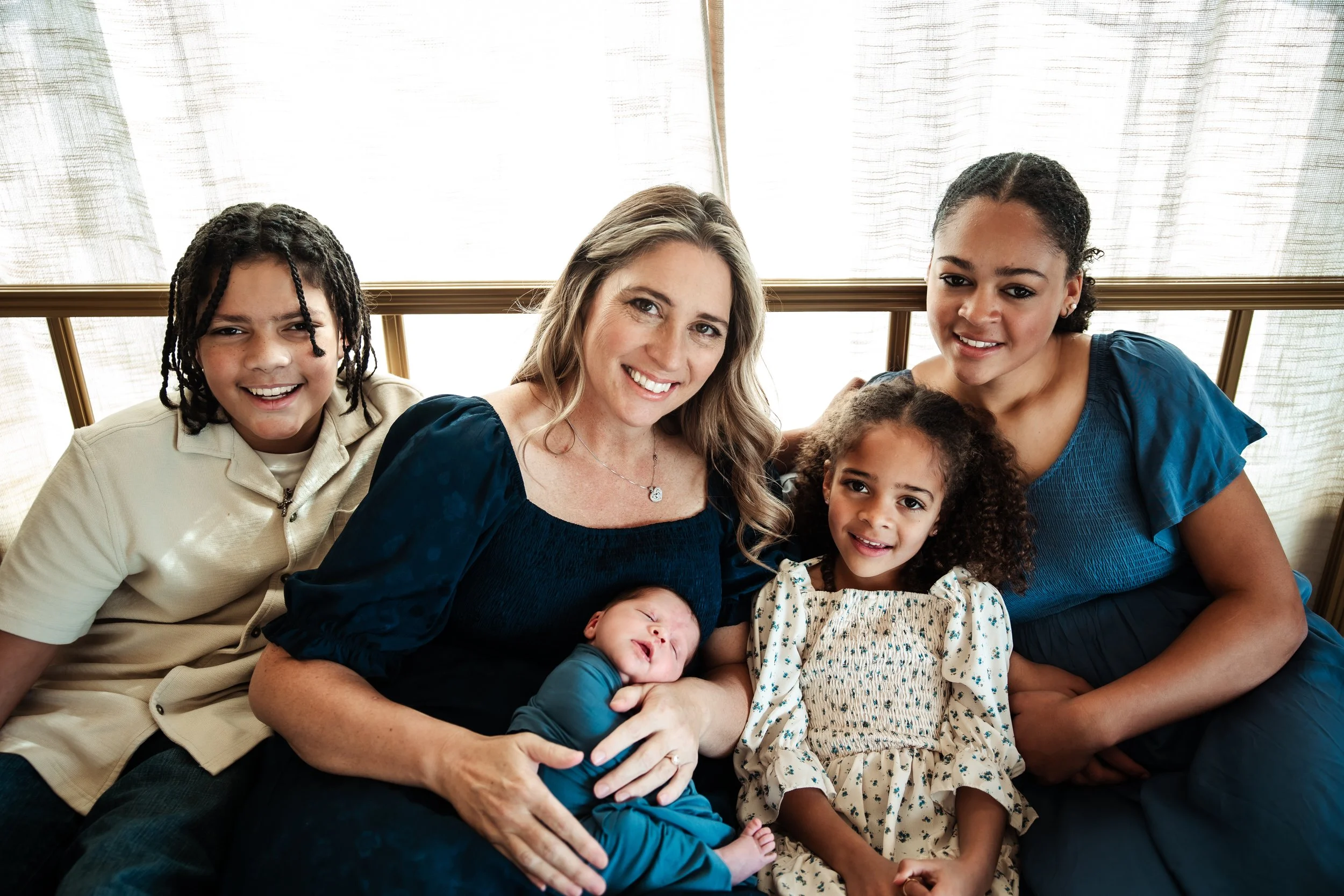 A woman sitting with four children, smiling at the camera, with a baby in her arms, seated between two young girls and a boy, in front of a window with light curtains.