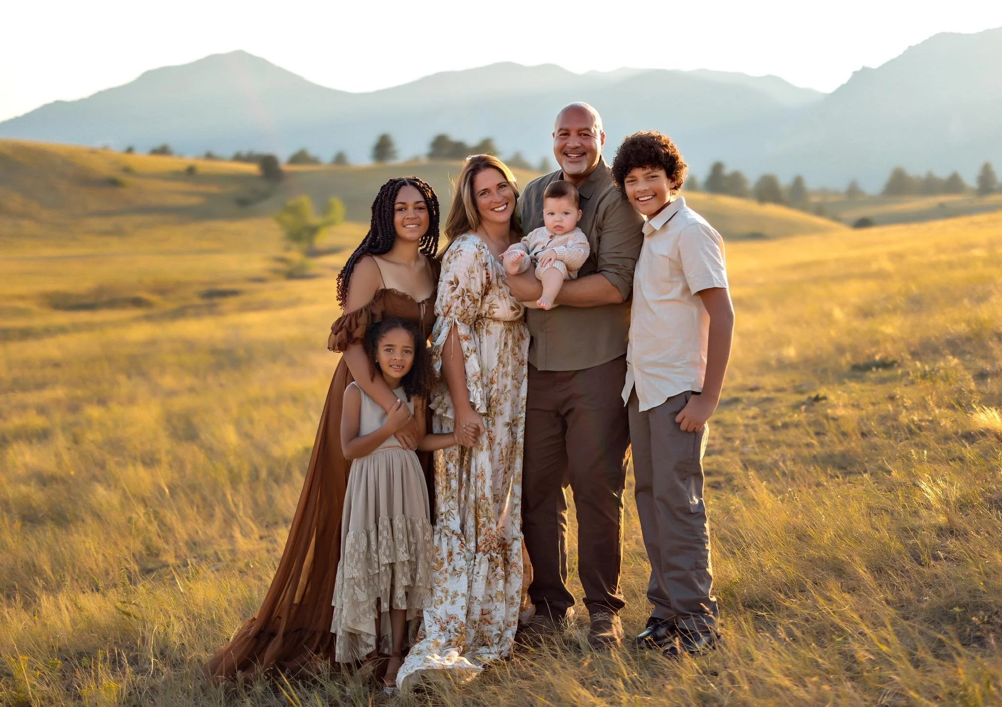 A family of seven standing in a grassy field with mountains in the background during sunset.