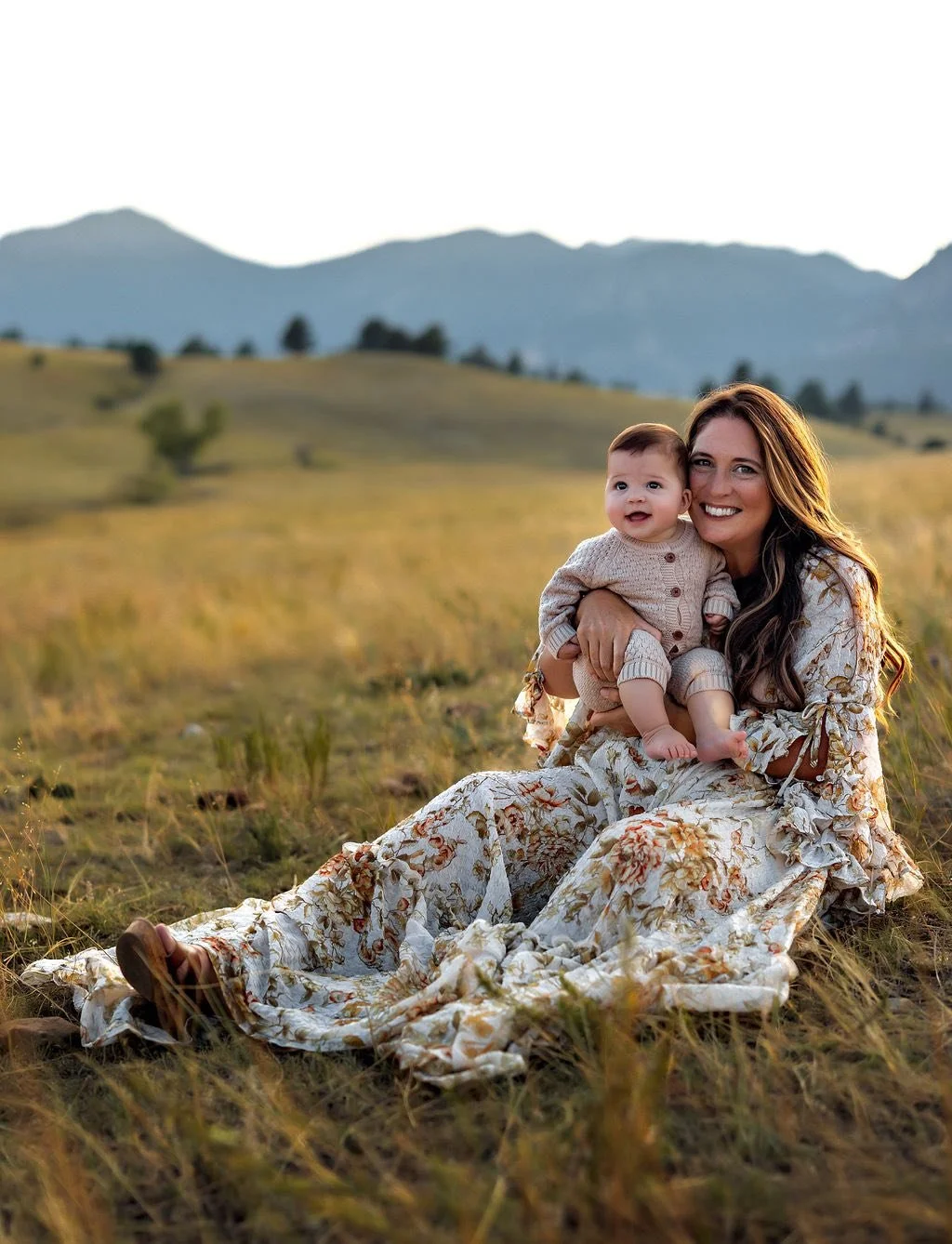 A woman with long brown hair smiling while sitting on grass in a floral dress, holding a baby in a beige knit outfit, with a scenic mountain landscape in the background.