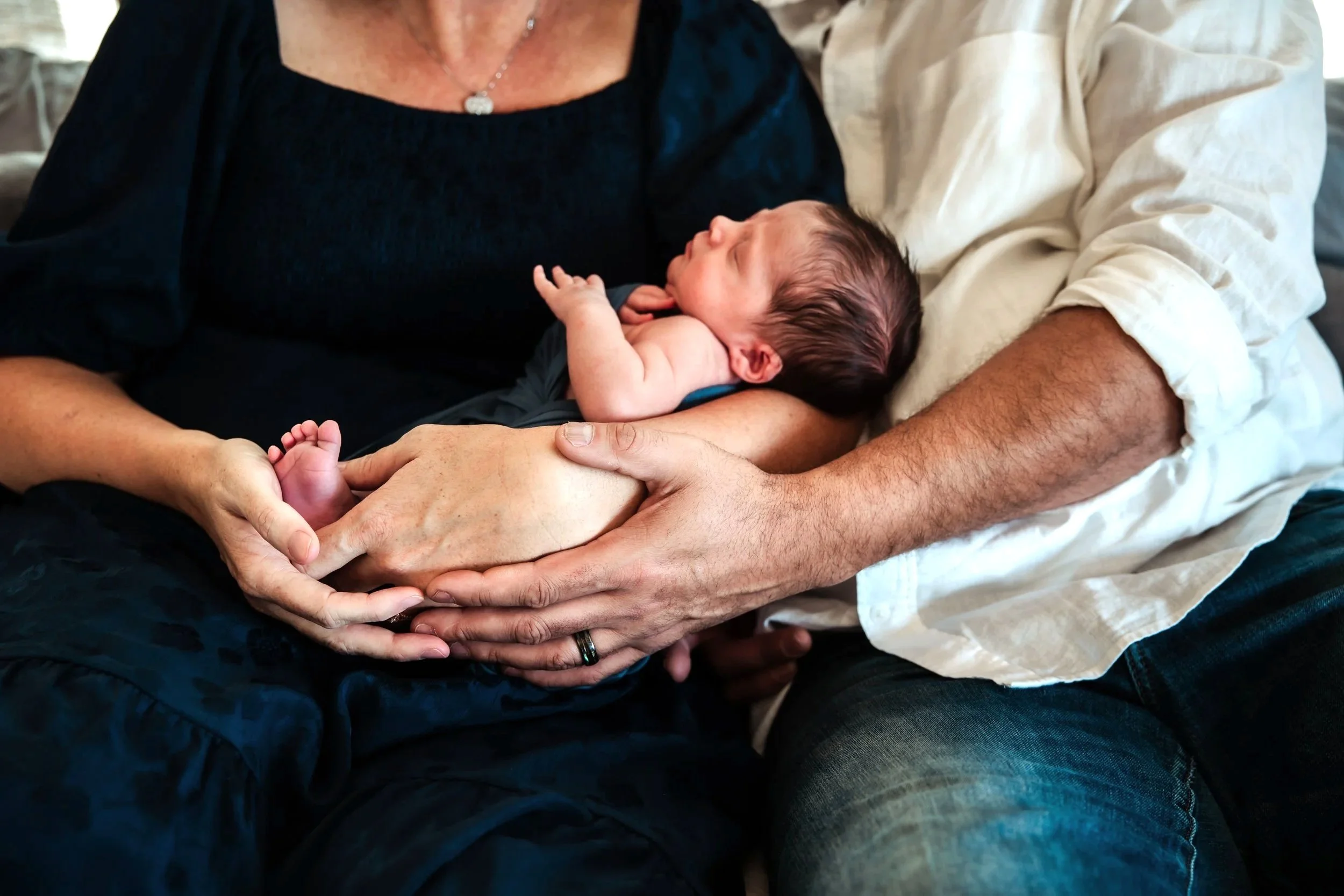A newborn baby resting on an older woman's chest, with the baby's hand touching the woman's arm. The man beside them is gently holding the baby's bottom, supporting the baby in a loving, caring pose.
