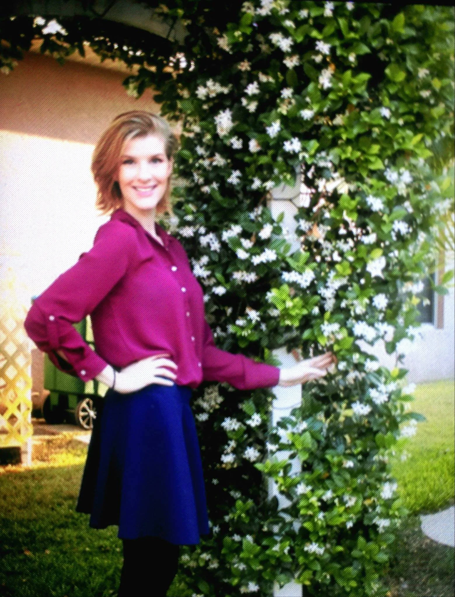 A woman in a purple shirt and black skirt standing outdoors next to a lush green hedge with white flowers, smiling at the camera.