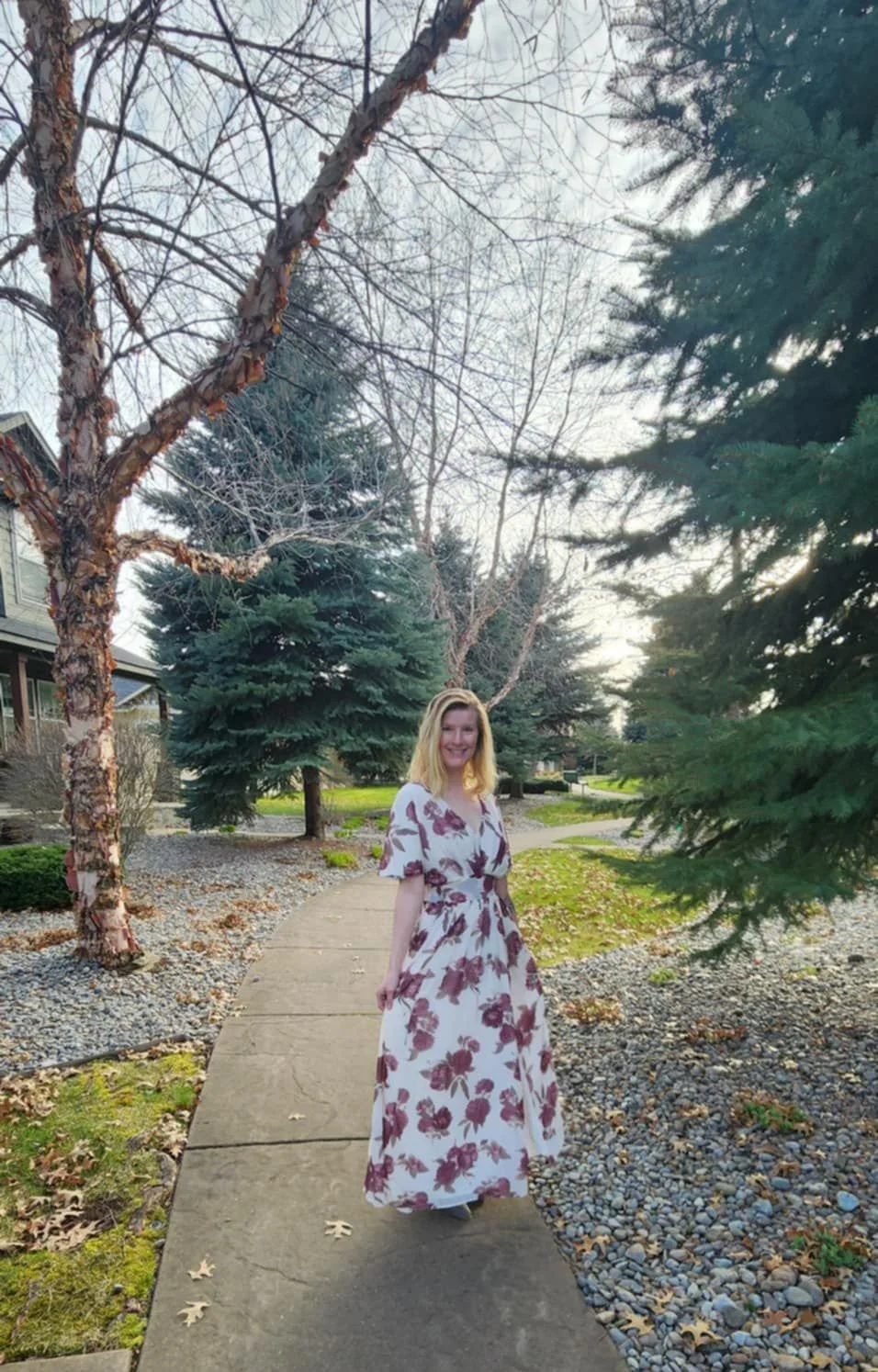 A woman in a white and purple floral dress standing on a sidewalk in a suburban neighborhood, surrounded by evergreen trees and leafless trees, during daytime.