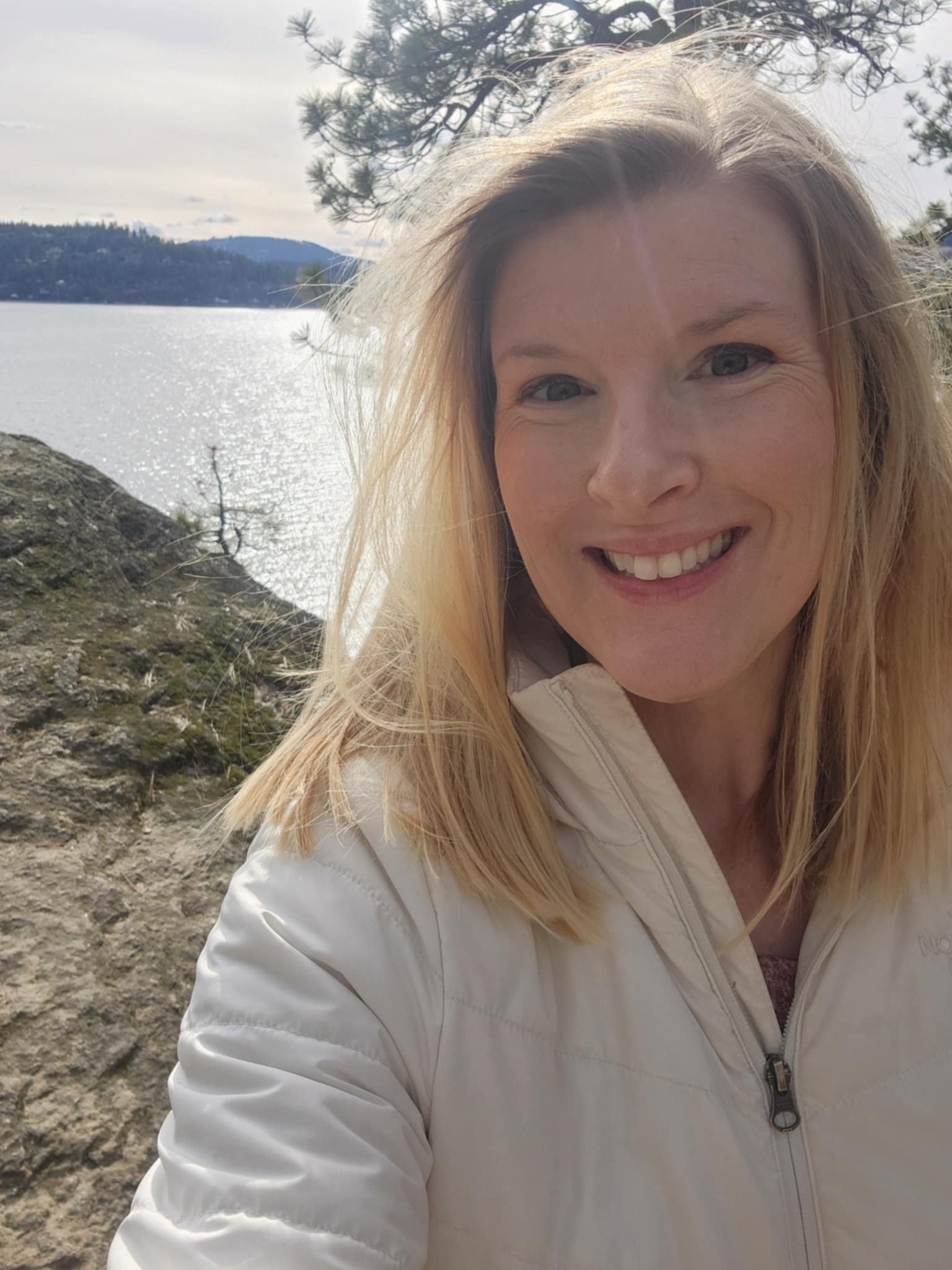 A woman with light blonde hair smiling outdoors near a lake, with trees and mountains in the background.