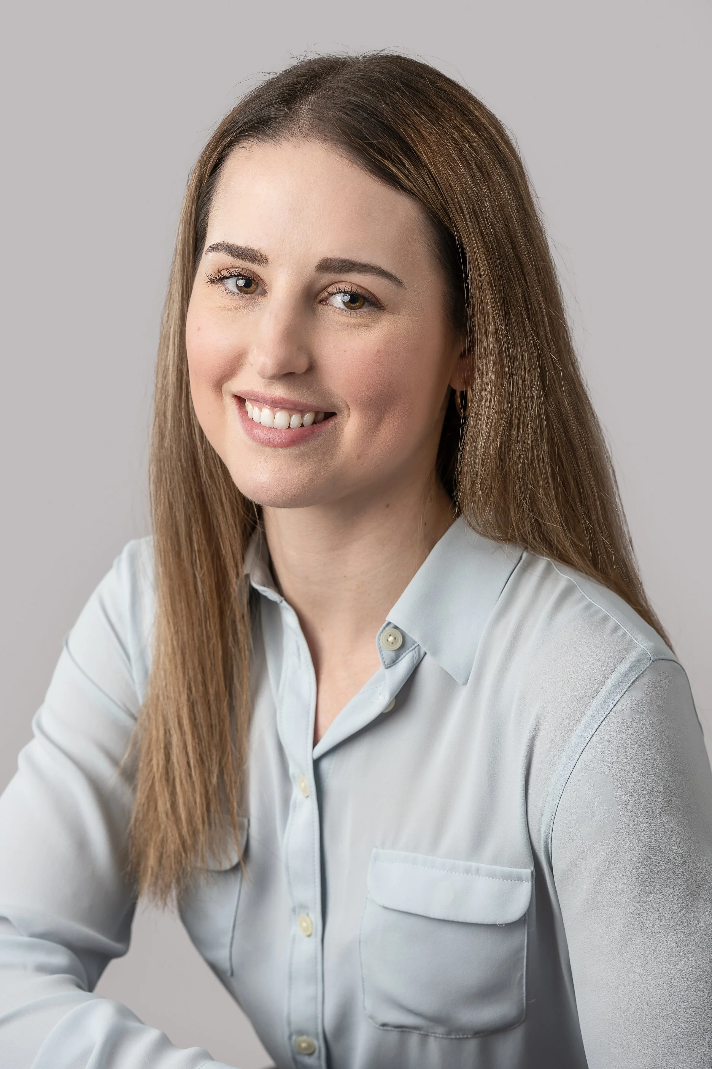 A young woman with long brown hair and light skin, wearing a light gray button-up shirt, smiling at the camera against a plain light gray background.