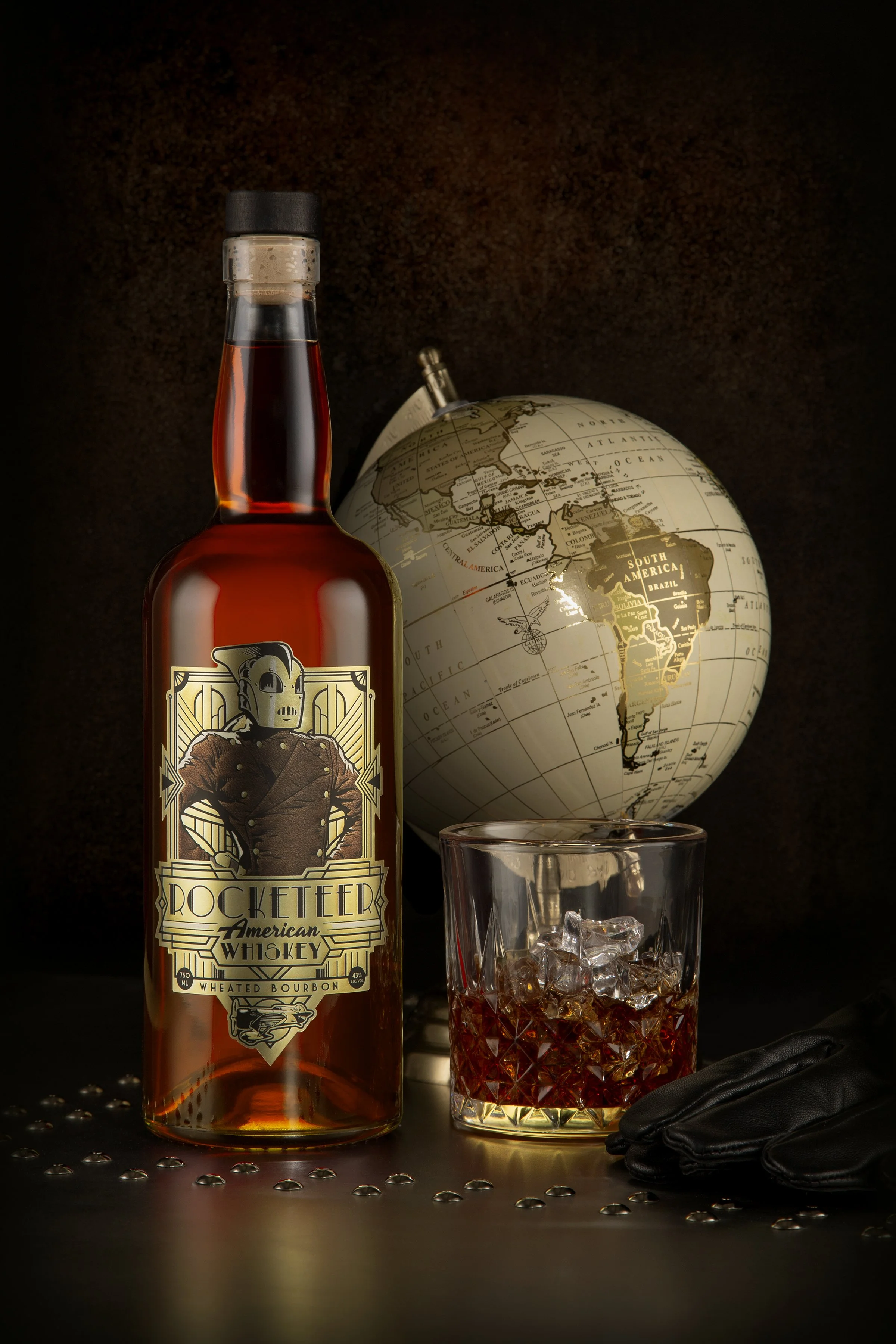 A bottle of Rocketter American whiskey, a glass with whiskey and ice, a black glove, a globe showing South America, and scattered silver dots on a dark surface.