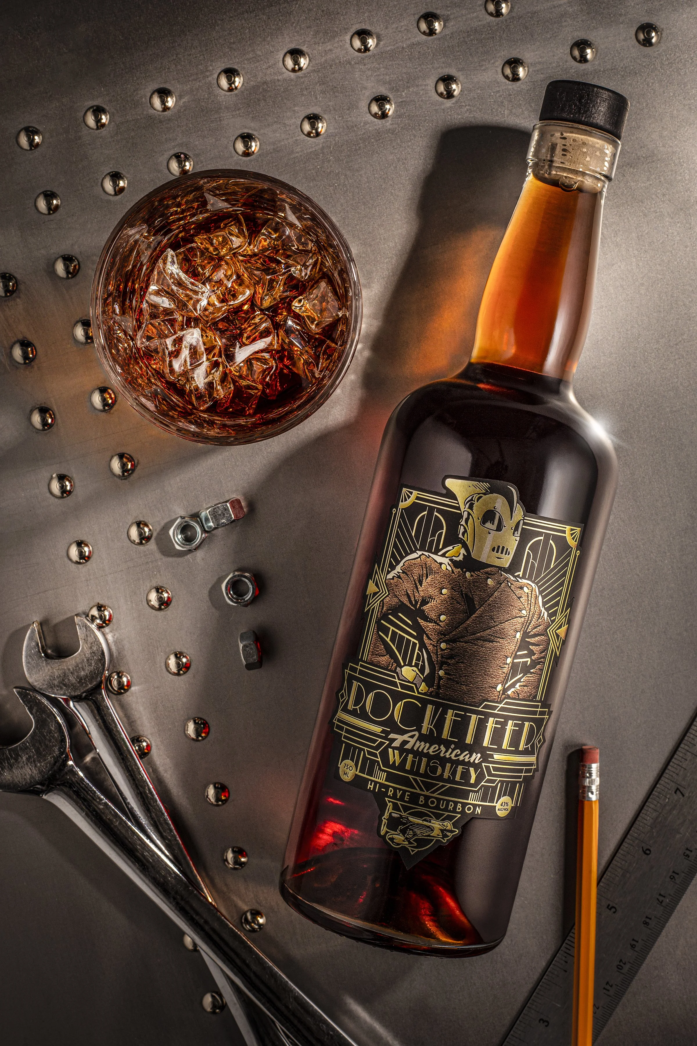 A bottle of Rockettier American Whiskey next to a glass of whiskey with ice, accompanied by tools and a ruler on a metal surface.