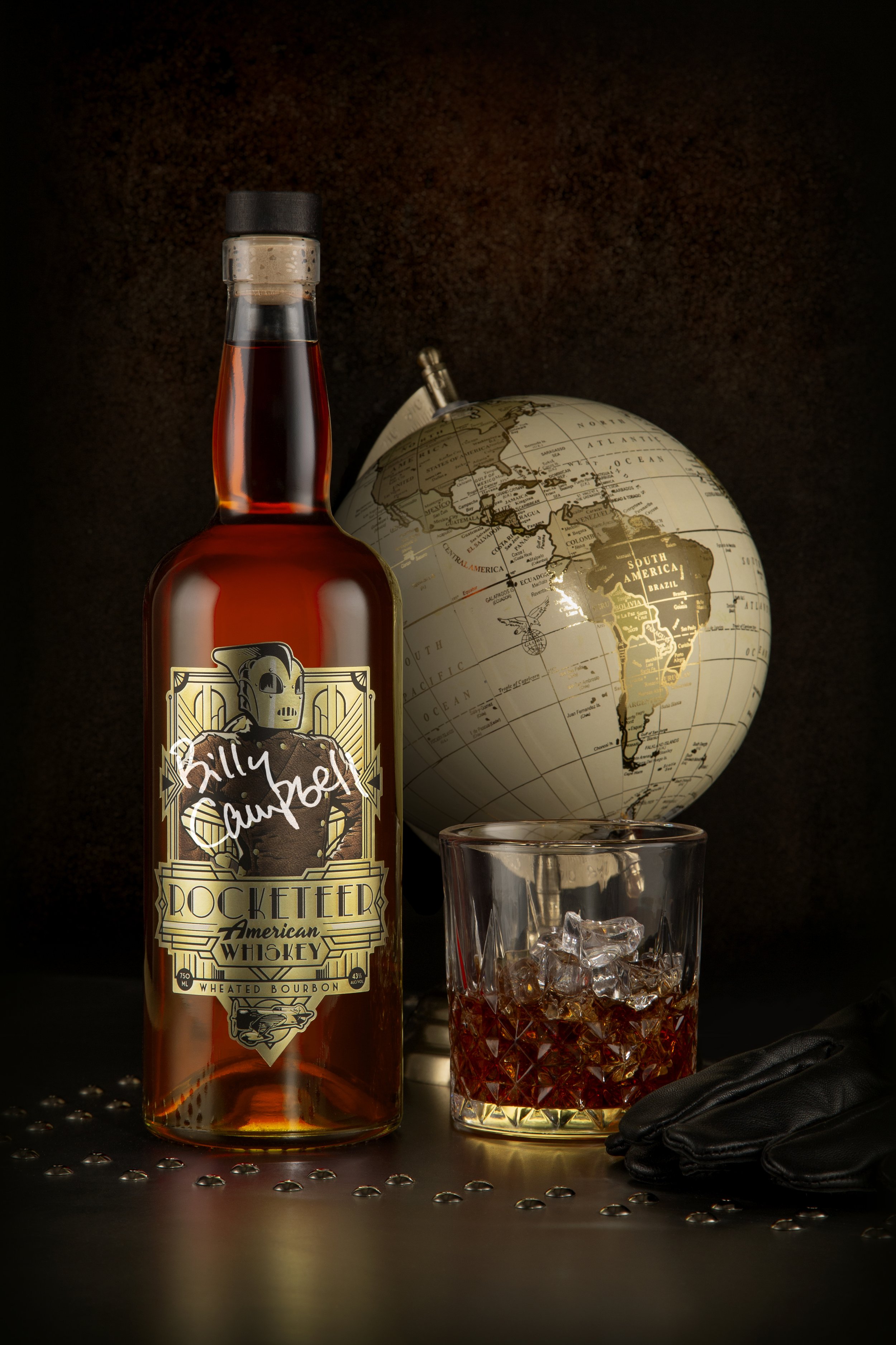 A bottle of Billy Campbell Rocketeer American Whiskey in front of a globe, with a glass of whiskey and ice, a black glove, and a metal surface with small round decorations.