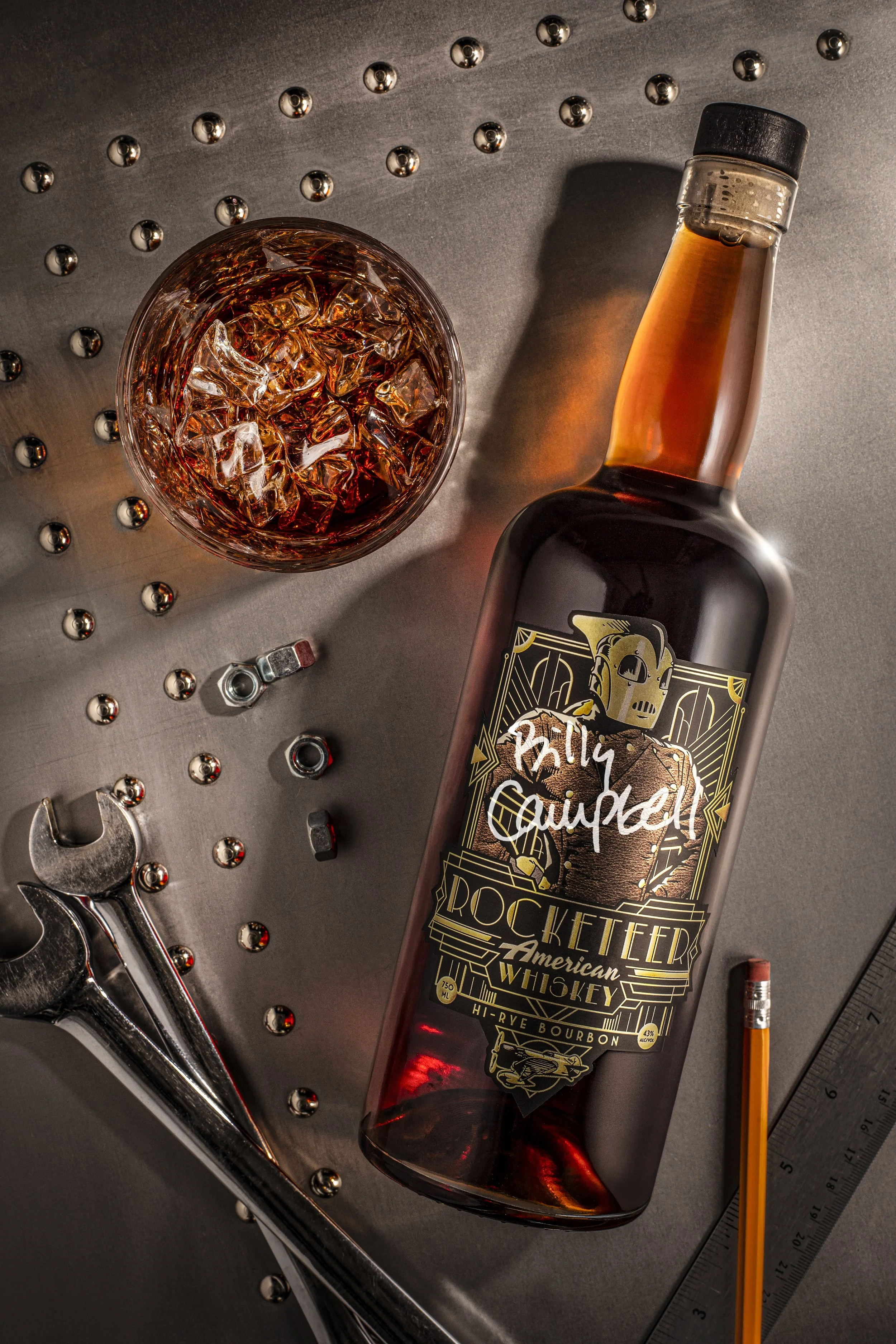 A bottle of rocketeer American whiskey with a black cap, a glass of whiskey with ice, a measuring ruler, a pencil, an open wrench, metal bolts, and metal balls on a metallic surface.