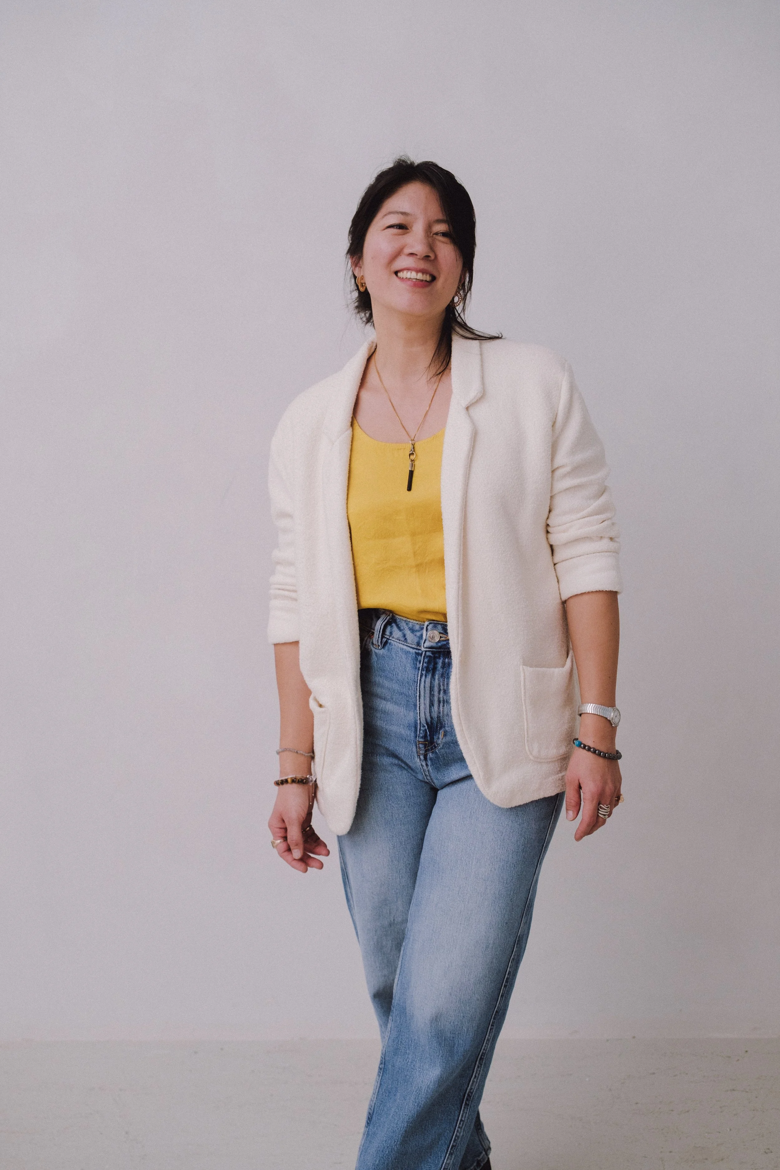 Stephanie Chung Reiki Master standing against a plain grey background, smiling, wearing a cream-colored blazer, a yellow top, and blue jeans, accessorised with jewellery and a watch.