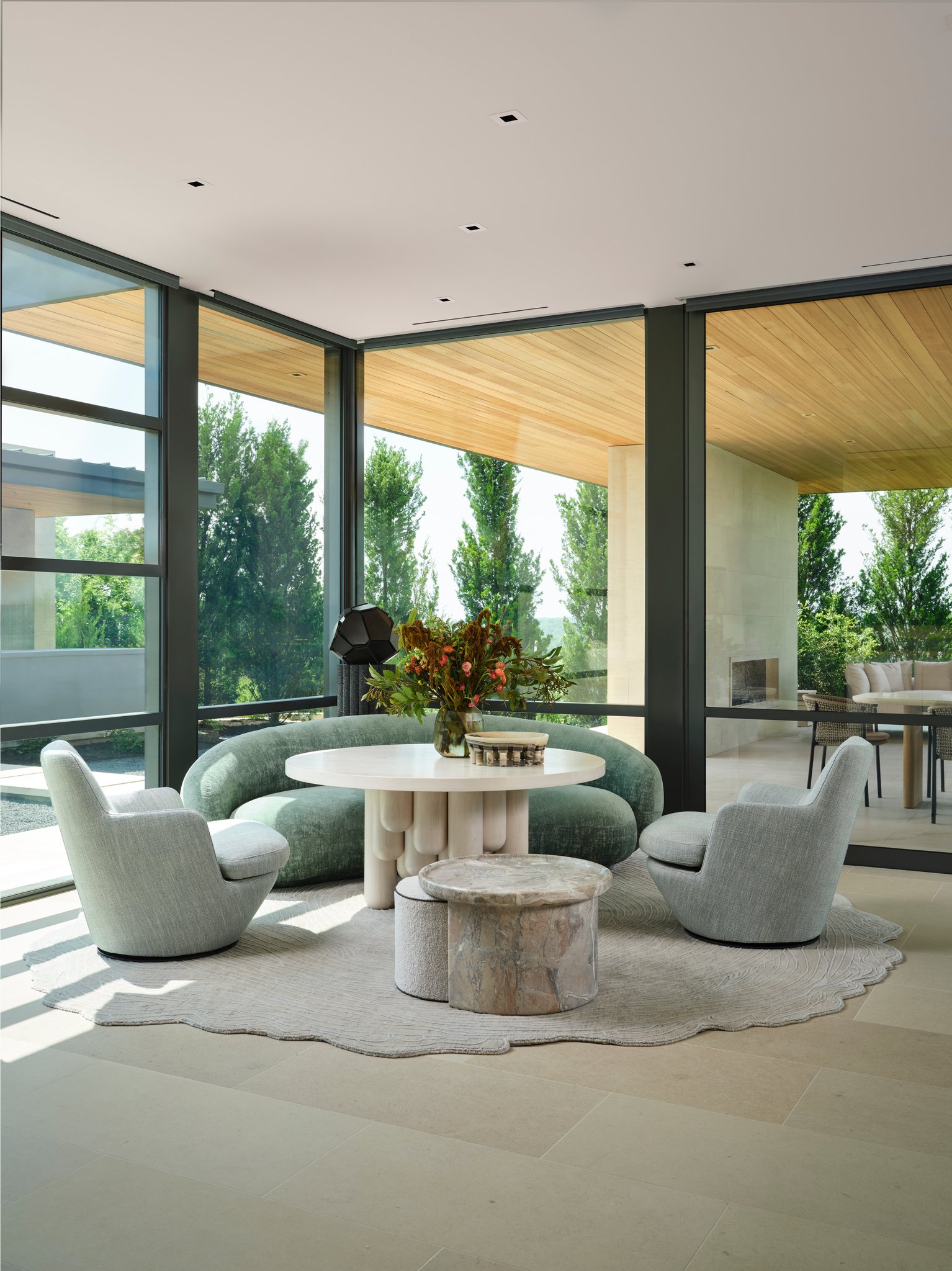 Modern breakfast area with curved sofa, sculptural round table, and floor-to-ceiling glass overlooking landscape. tacchini sofa, rische collection seren table arik levy sculpture, bensen chairs, atelier agahzadeh rug