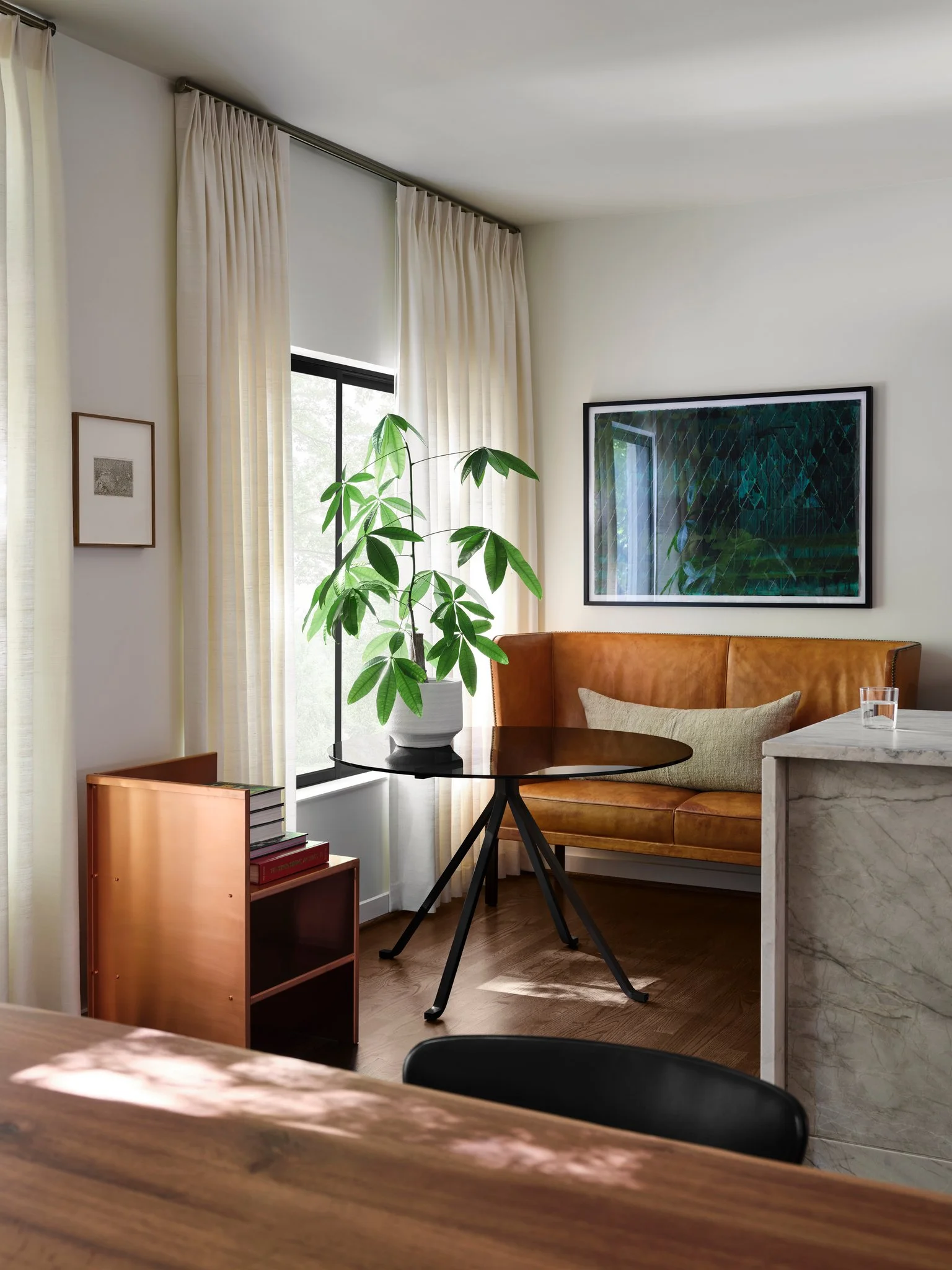 Modern breakfast nook with leather sofa by Frits Henningsen, round glass table by Enzo Mari and linen drapery, set against warm wood flooring and natural light. Copper chair by Donald Judd in the foreground.