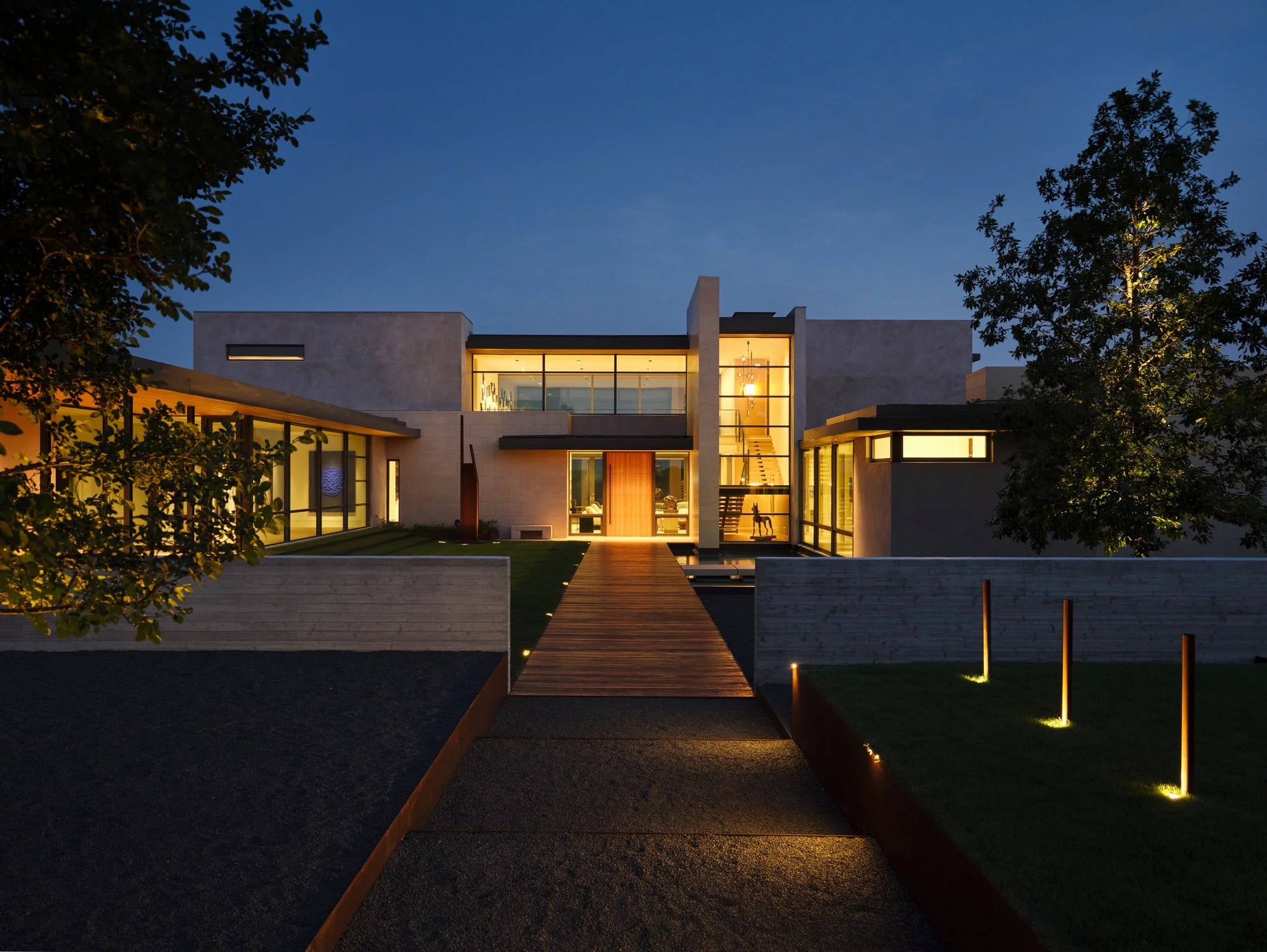 Evening entry approach view of a contemporary home with illuminated glass stair hall, limestone volumes, and a long wood entry bridge crossing a reflecting water feature.