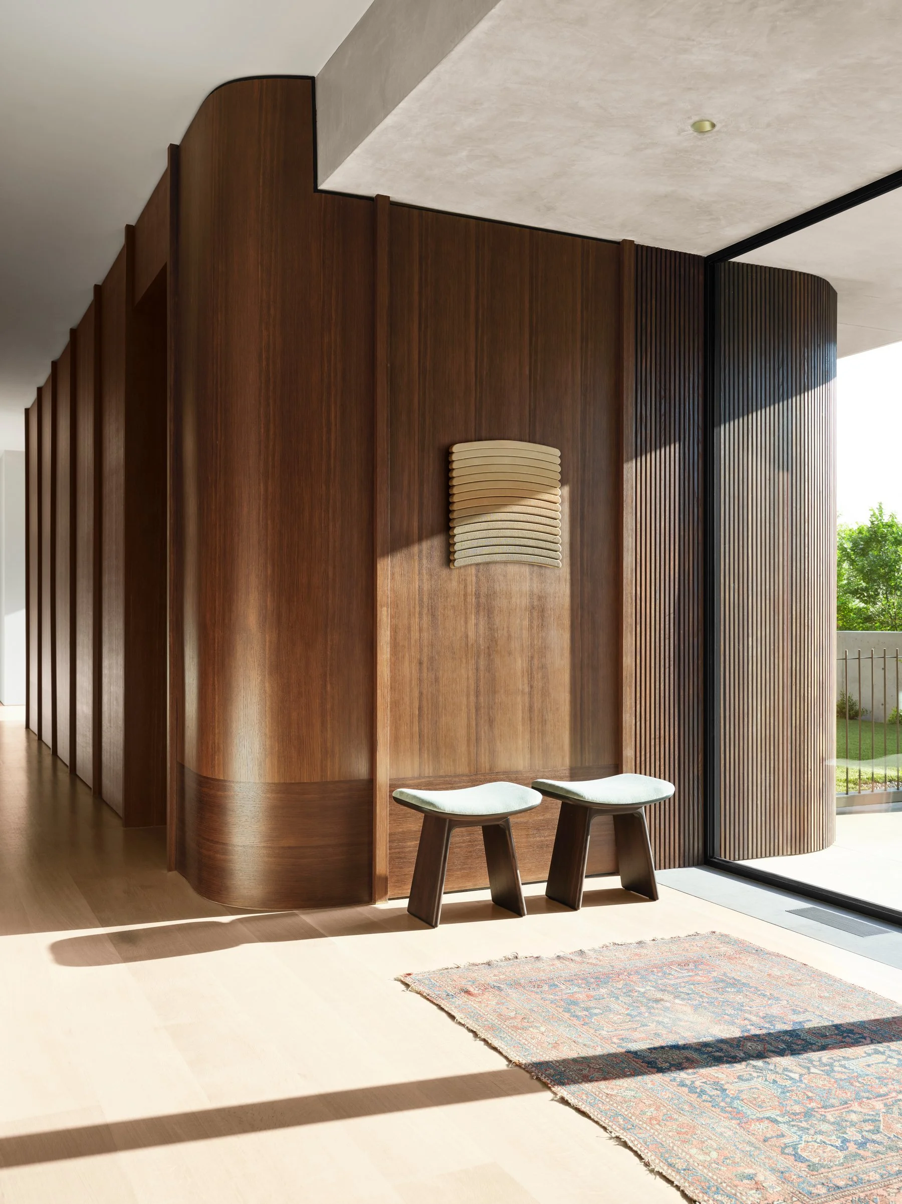 Curved fumed white oak wood-paneled wall with custom millwork and sculptural stools in a contemporary residential interior with natural light and warm materials. Art on wall by Luke Diiorio