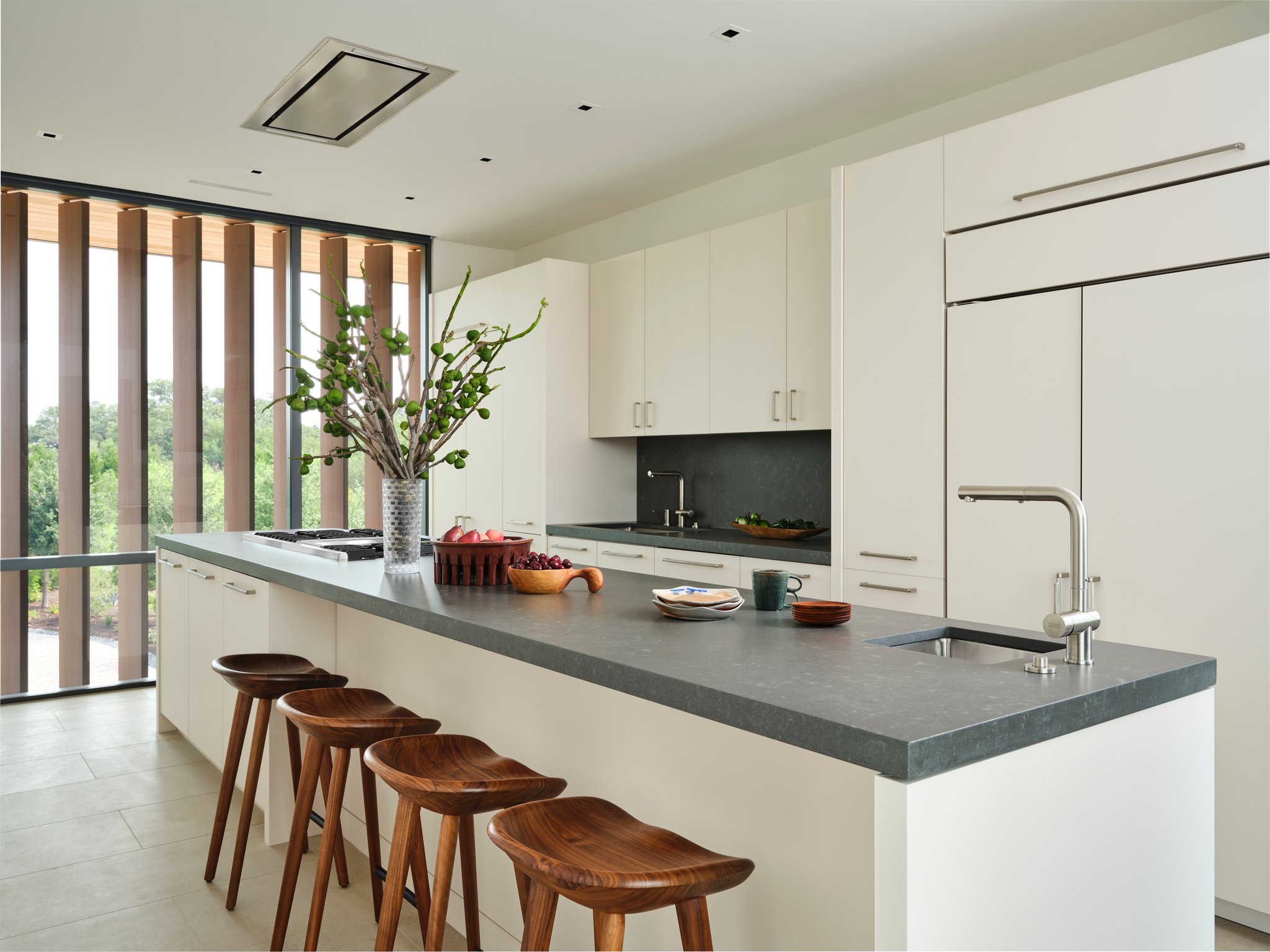 Minimal kitchen with large stone island, white cabinetry, and sculptural walnut bar stools. Floor-to-ceiling glazing frames landscape views and brings natural light into the space. bassam fellows
