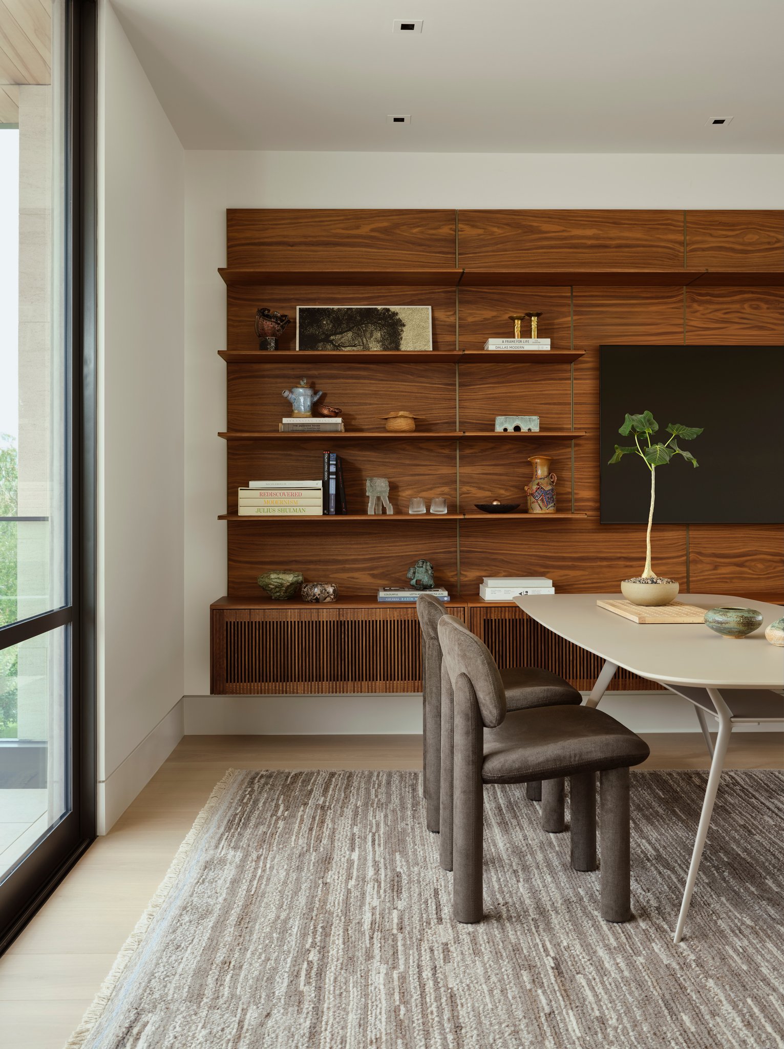 Home office framed by custom walnut shelving displaying books, ceramics, and objects. Warm wood paneling contrasts with light flooring and sculptural dining furniture.