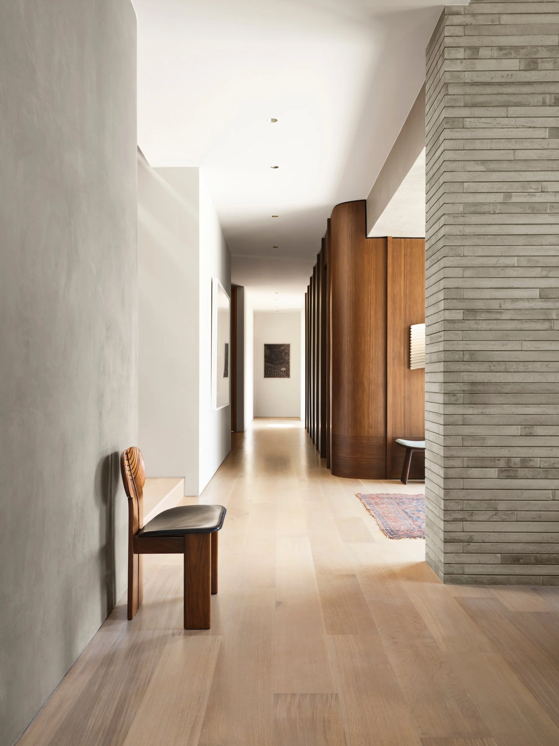 Linear corridor with plaster walls, wood paneling, and integrated seating, establishing rhythm and spatial continuity. Vintage Tobia and Afra Scarpa ‘Artona’ chair in foreground.