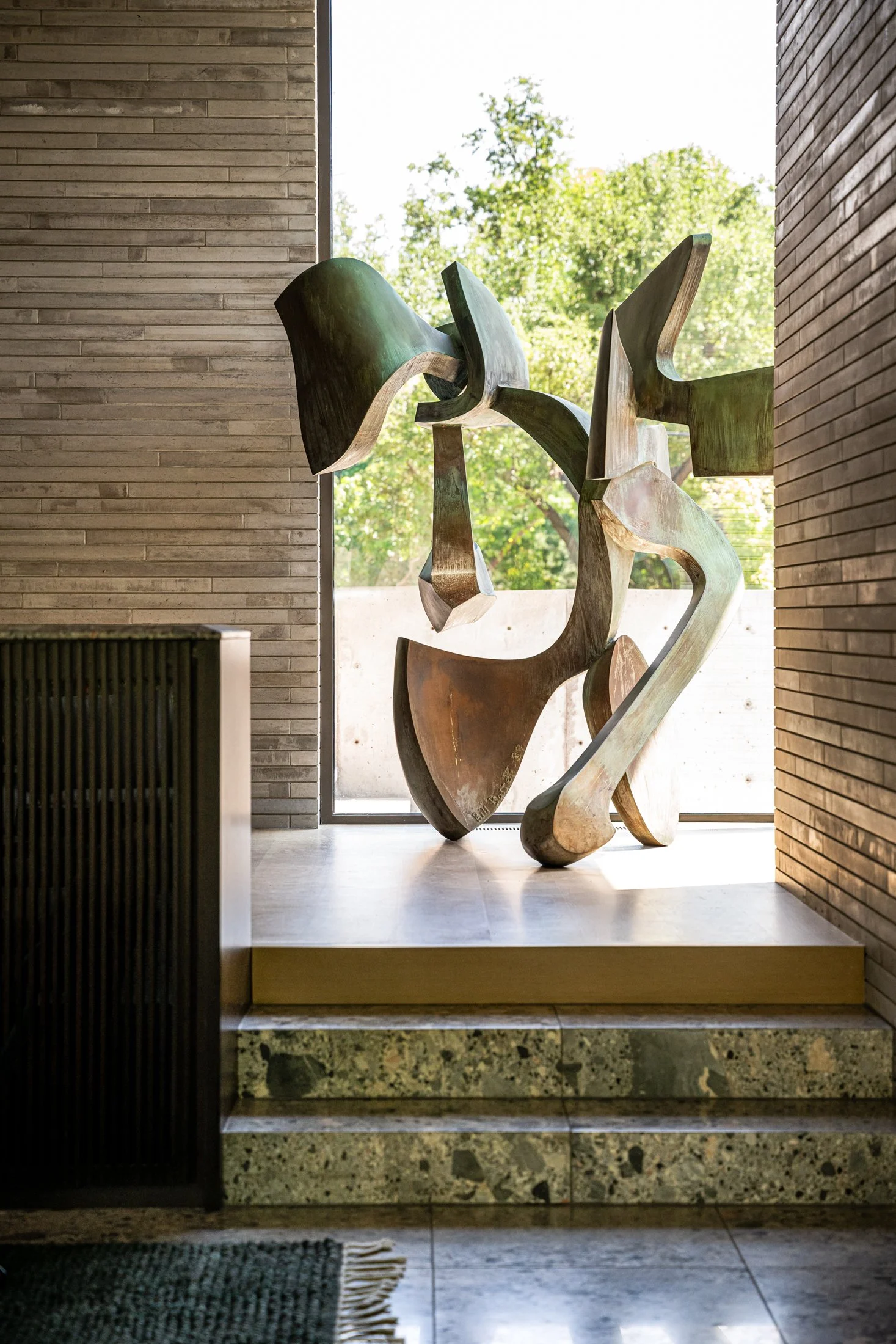 Large-scale abstract Bronze mid-century sculpture by Bill Barrett set within a light-filled modern interior, framed by brick walls and floor-to-ceiling glazing.