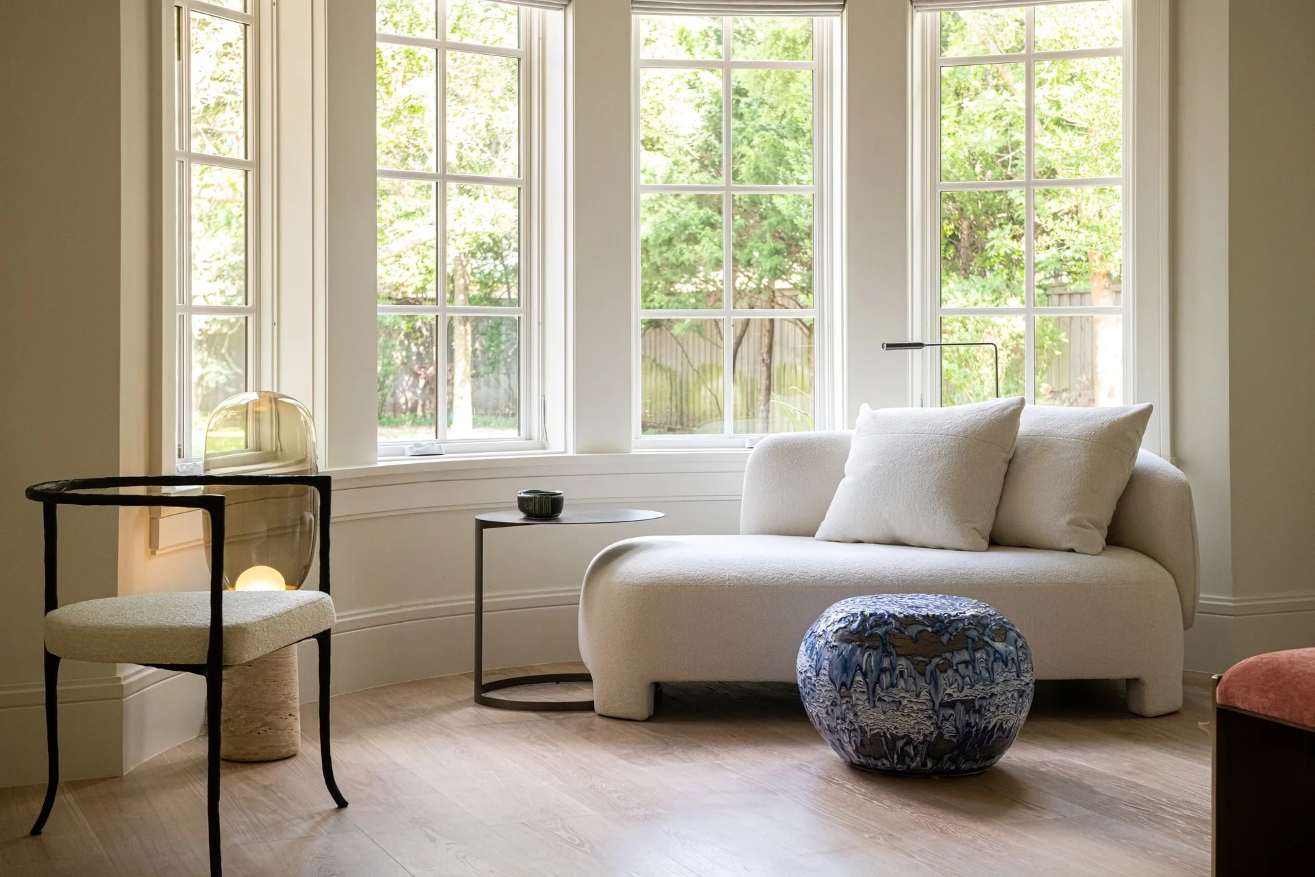 Bay window lounge with curved bouclé chaise by Sebastian Herkner, collectible ceramic stool by pierre yovanovitch , and sculptural bronze accent chair in a warm contemporary residential interior.