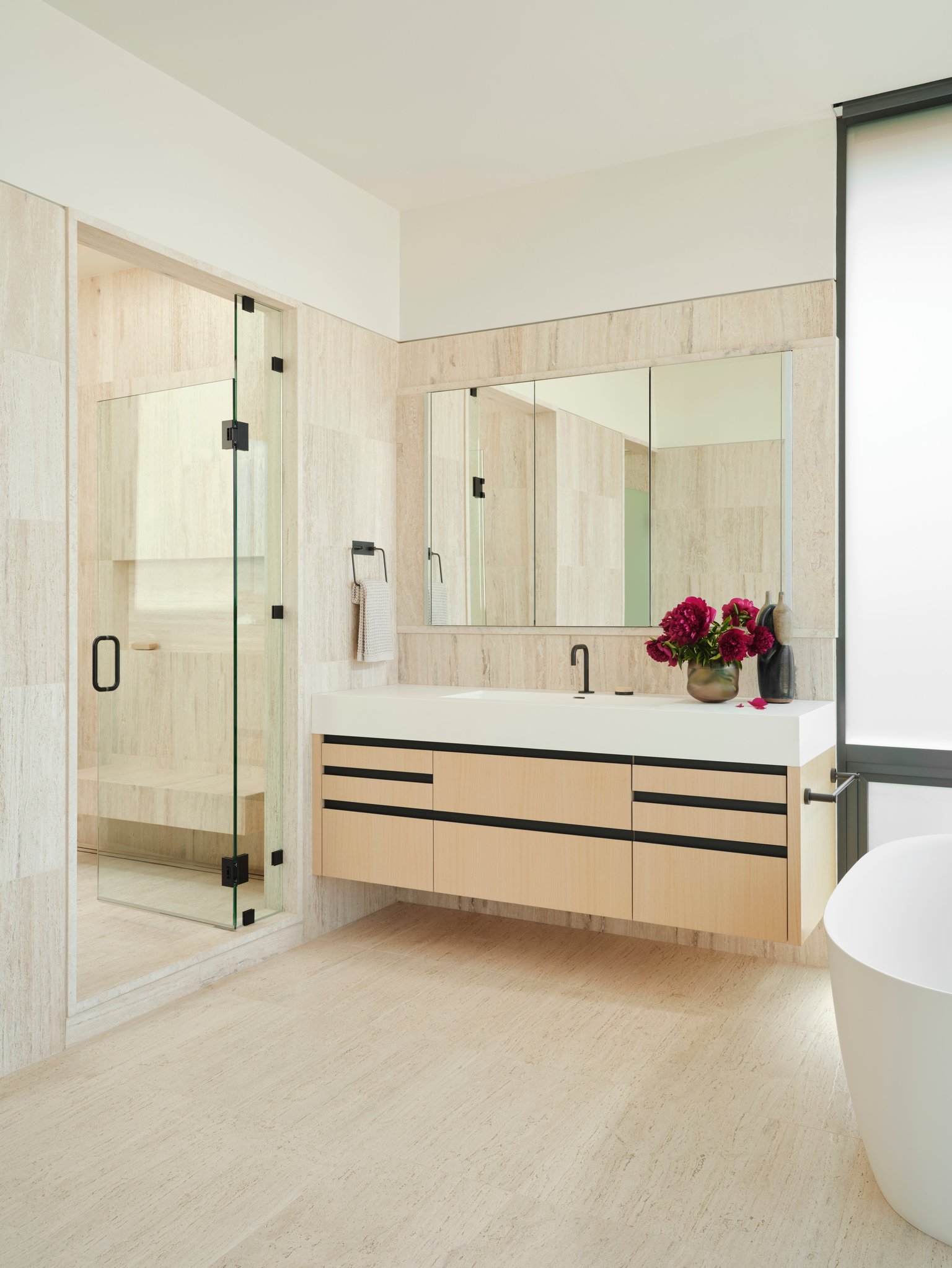 Light-filled bathroom clad in honed travertine with frameless glass shower, floating oak vanity, and restrained black fixtures. A calm modern interior emphasizing natural stone, clean lines, and quiet material palette.