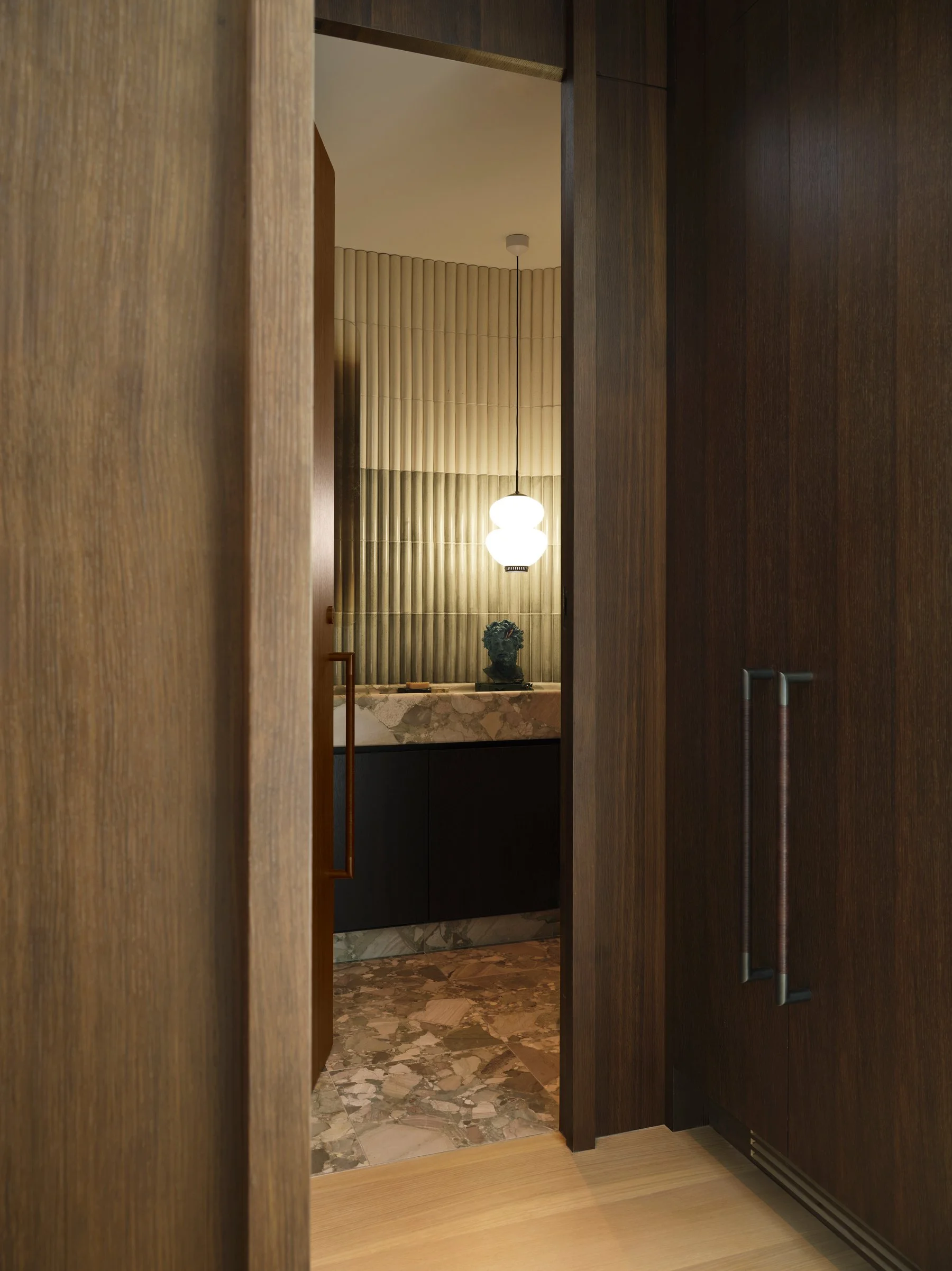 View through paneled passage into powder room, framing custom green ceppo marble vanity and pendant lighting within a controlled architectural sequence.