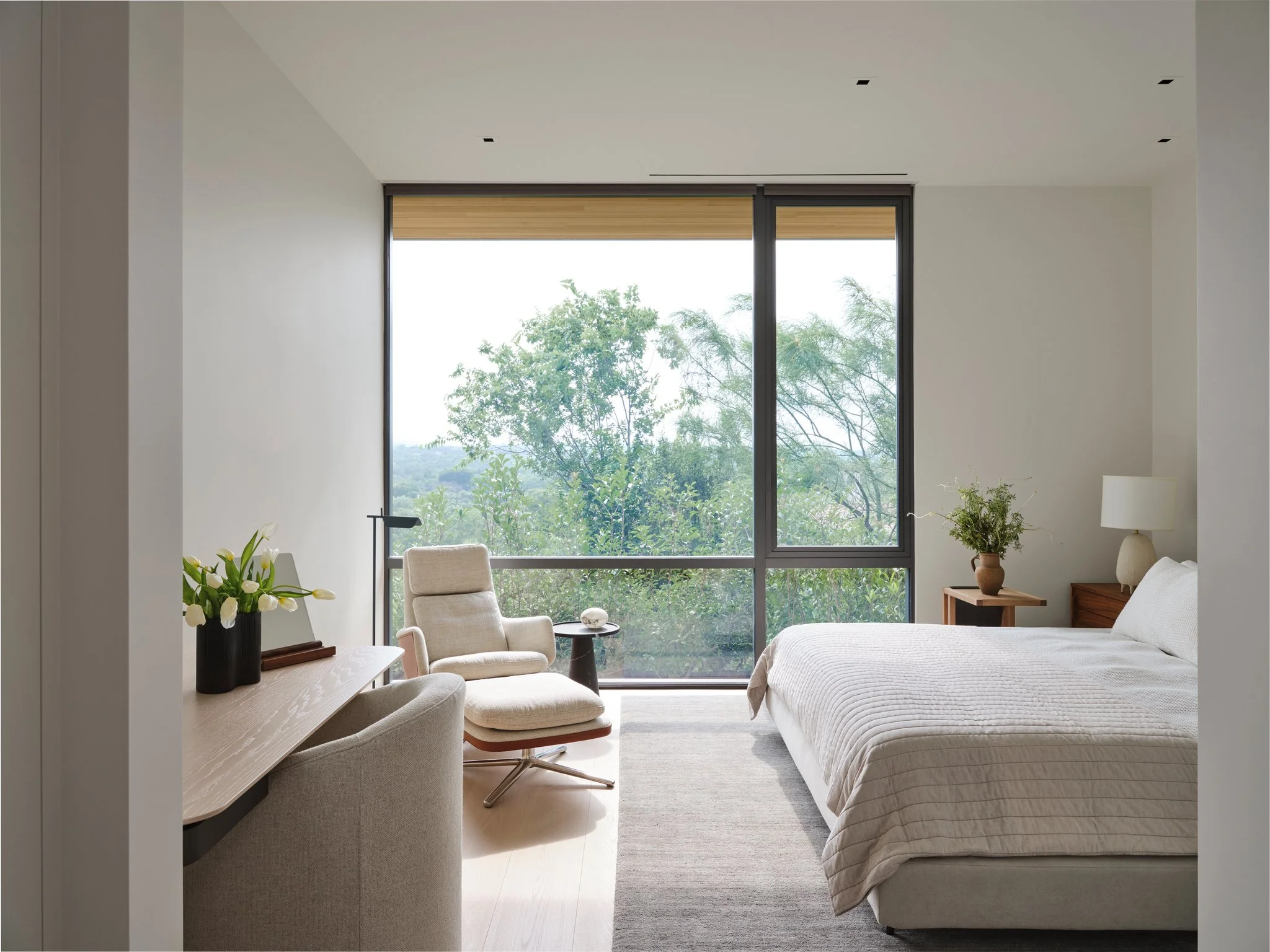 Minimal bedroom with soft neutral palette, lounge chair, and expansive window overlooking wooded landscape. Warm wood accents and natural light create a quiet retreat.