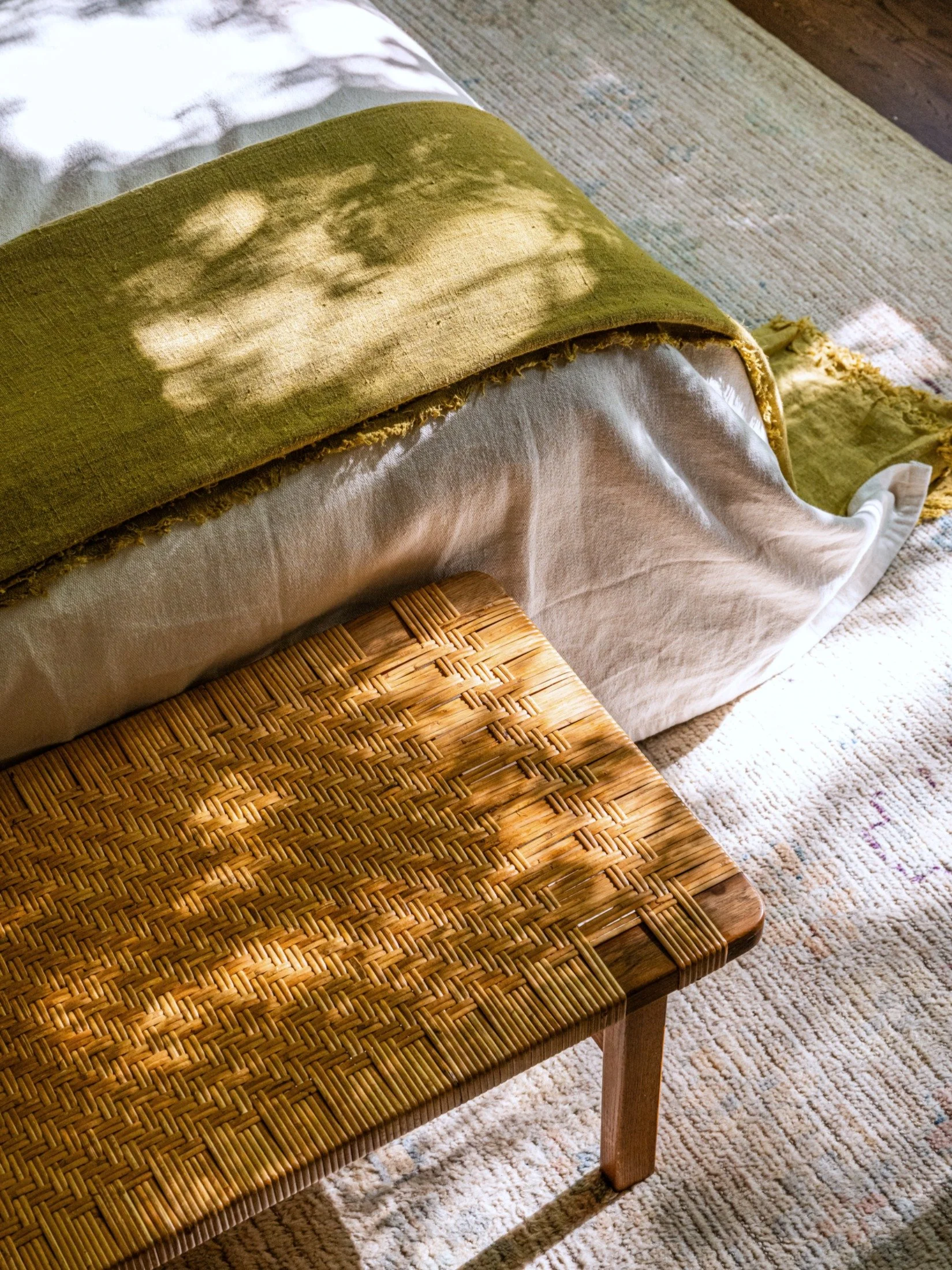 A woven cane bench by Bruno Mattheson at the foot of a bed dappled in natural light in a minimalist warm interior.