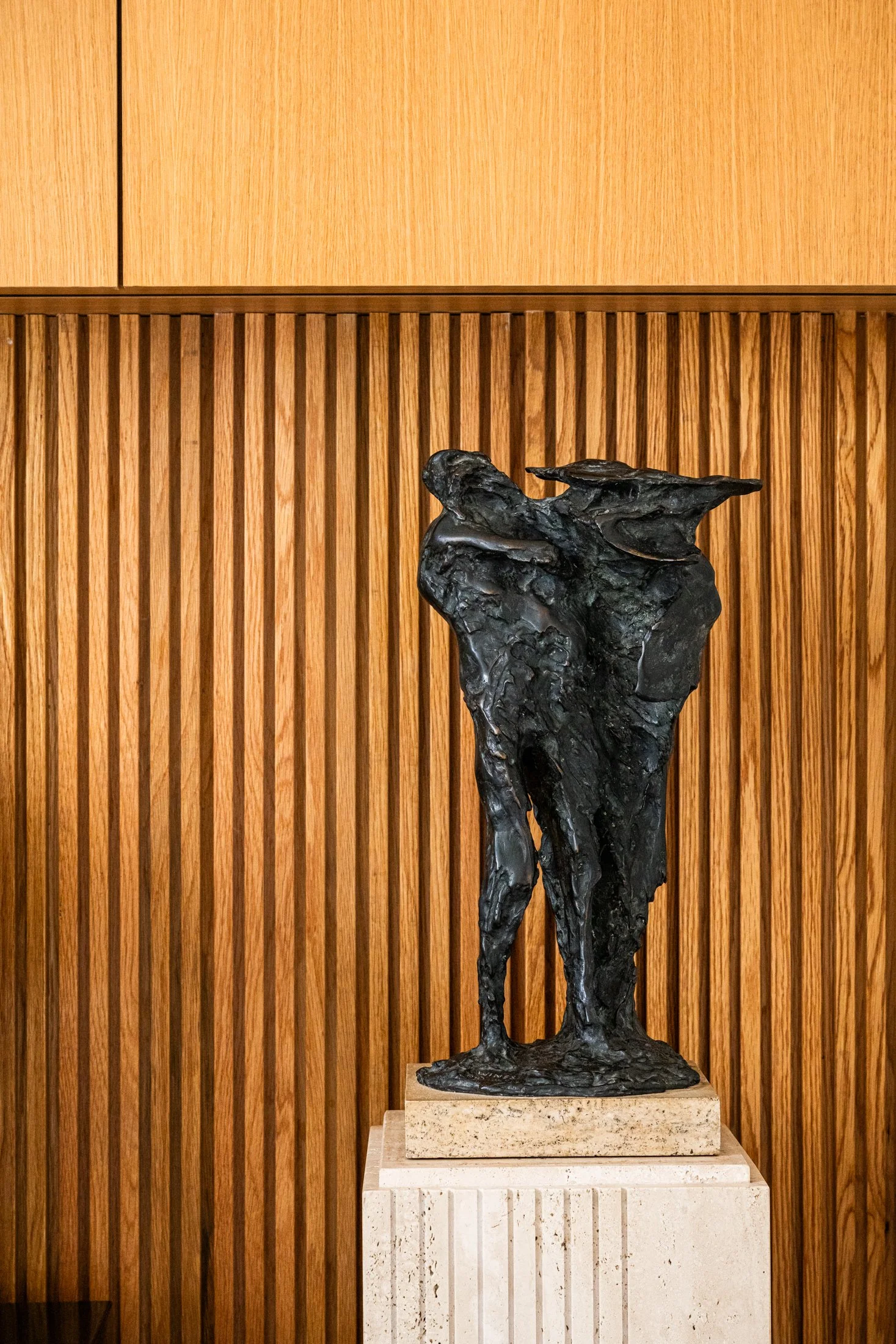 Bronze figurative sculpture on stone plinth set against vertical wood paneling, emphasizing contrast between organic form and linear backdrop.