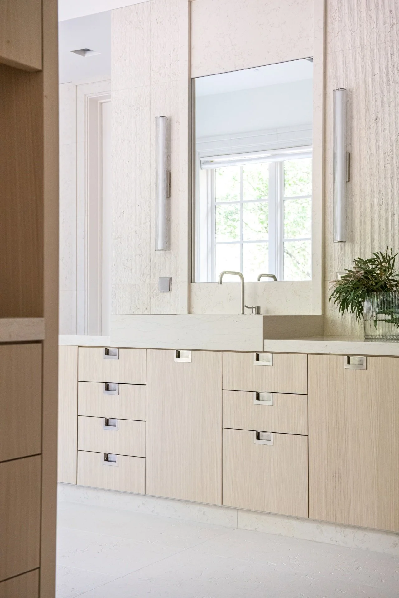 Primary bathroom vanity with custom pale wood cabinetry, integrated stone sink, textured limestone walls, and tailored modern detailing.
