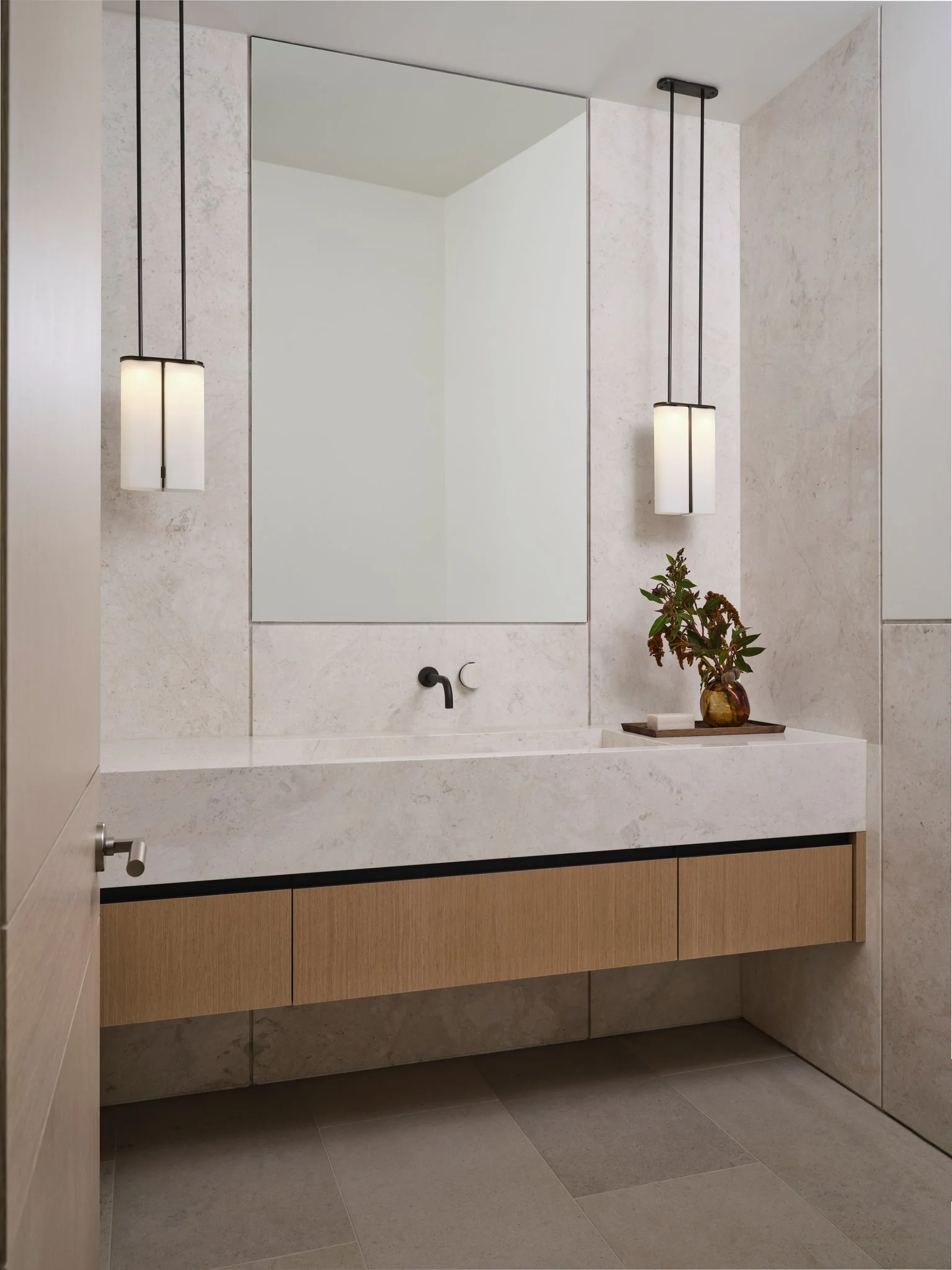 Powder room with floating oak vanity, integrated stone sink, and suspended pendant lights. Light stone walls and restrained detailing create a quiet, modern atmosphere.