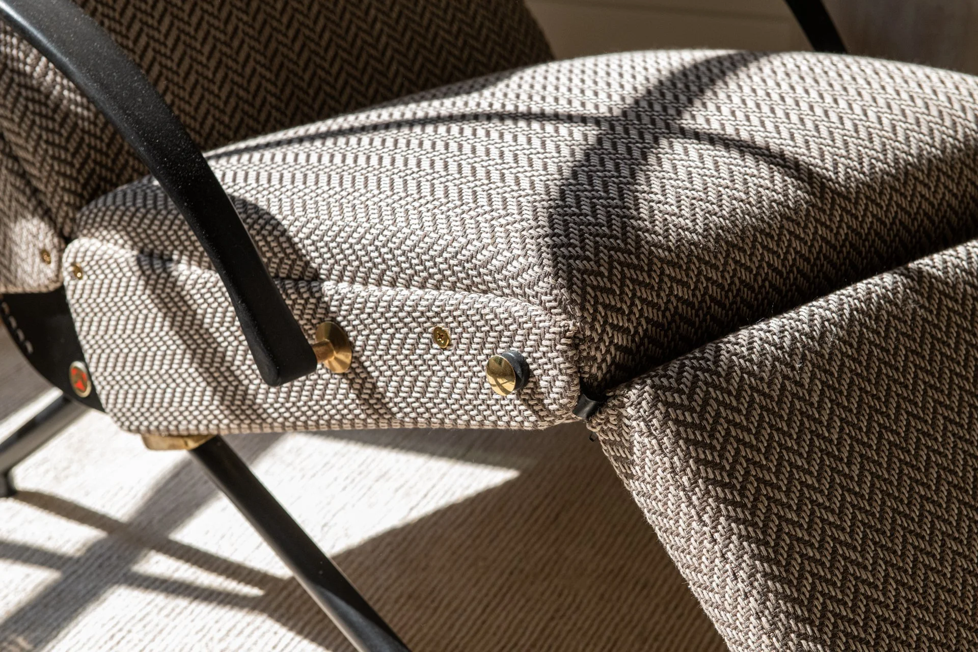 Close-up of tailored vintage lounge chair by Osvaldo Borsani upholstery with textured woven fabric, black steel frame, and brass detailing in dappled natural sunlight.