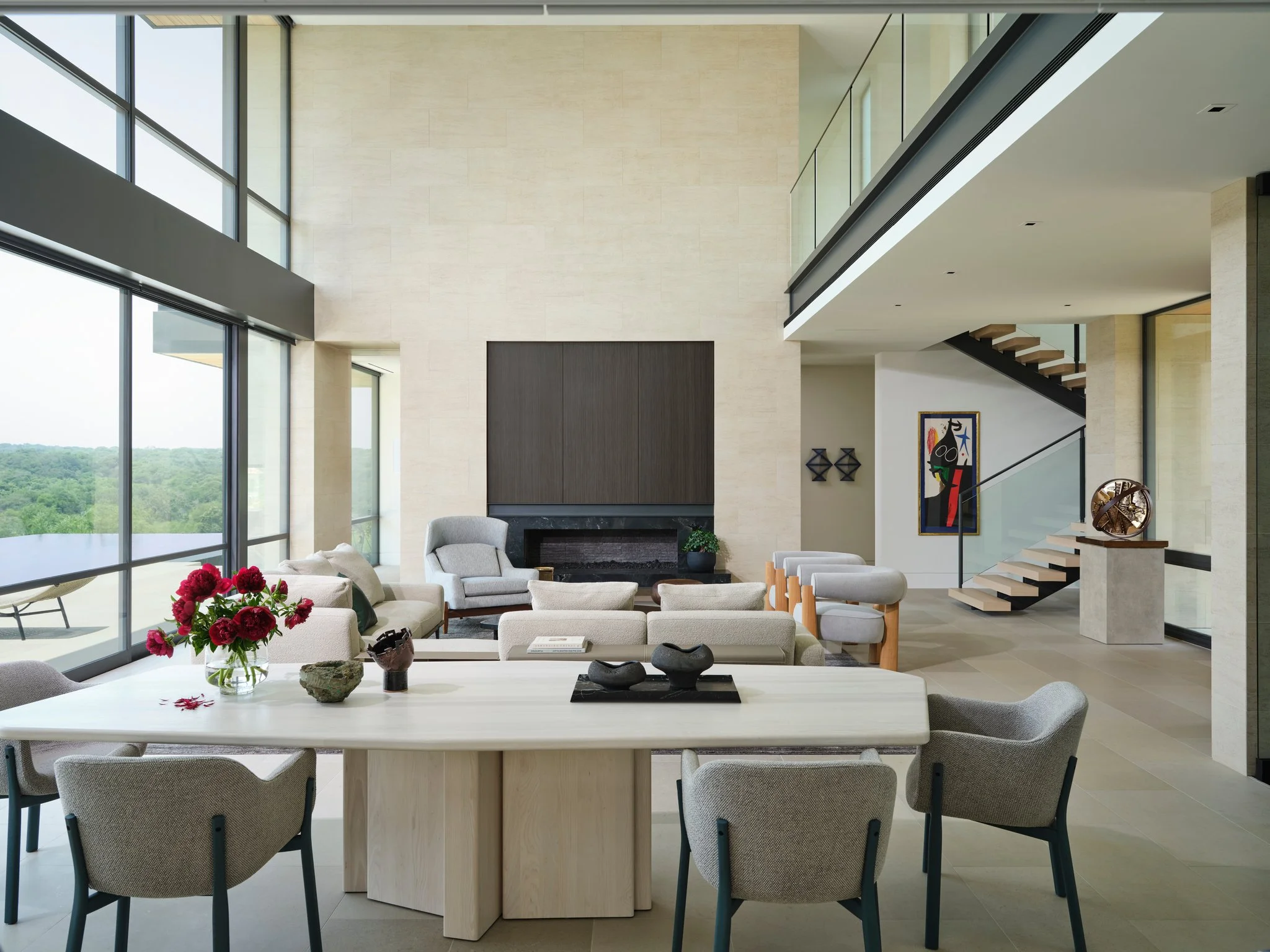 Open living and dining space with floor-to-ceiling glass, limestone walls, and floating stair. Sculptural furniture and curated artwork anchor the double-height interior. table by simon johns, poltrona frau, joan miro and arnaldo pomodoro sculpture